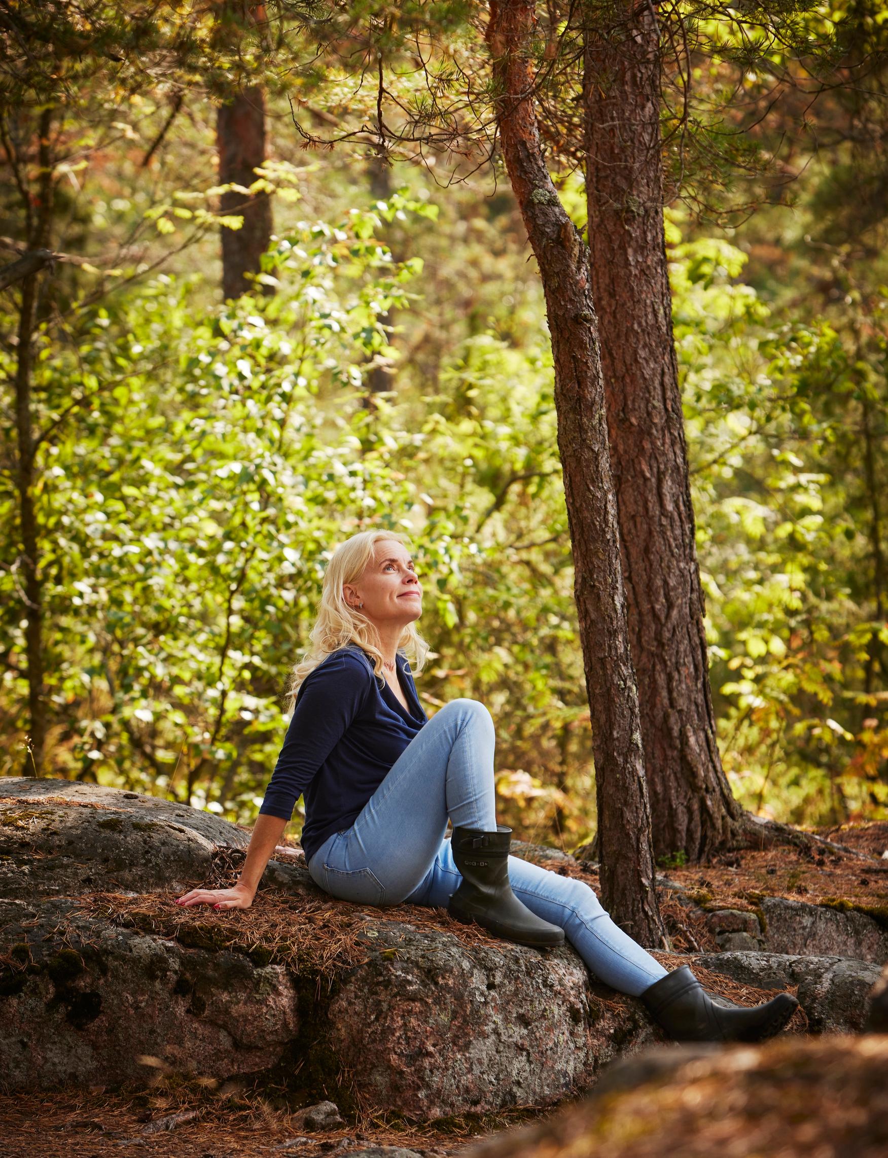 nainen istuu kalliolla, taustalla kasvaa mäntyjä ja lehtimetsää