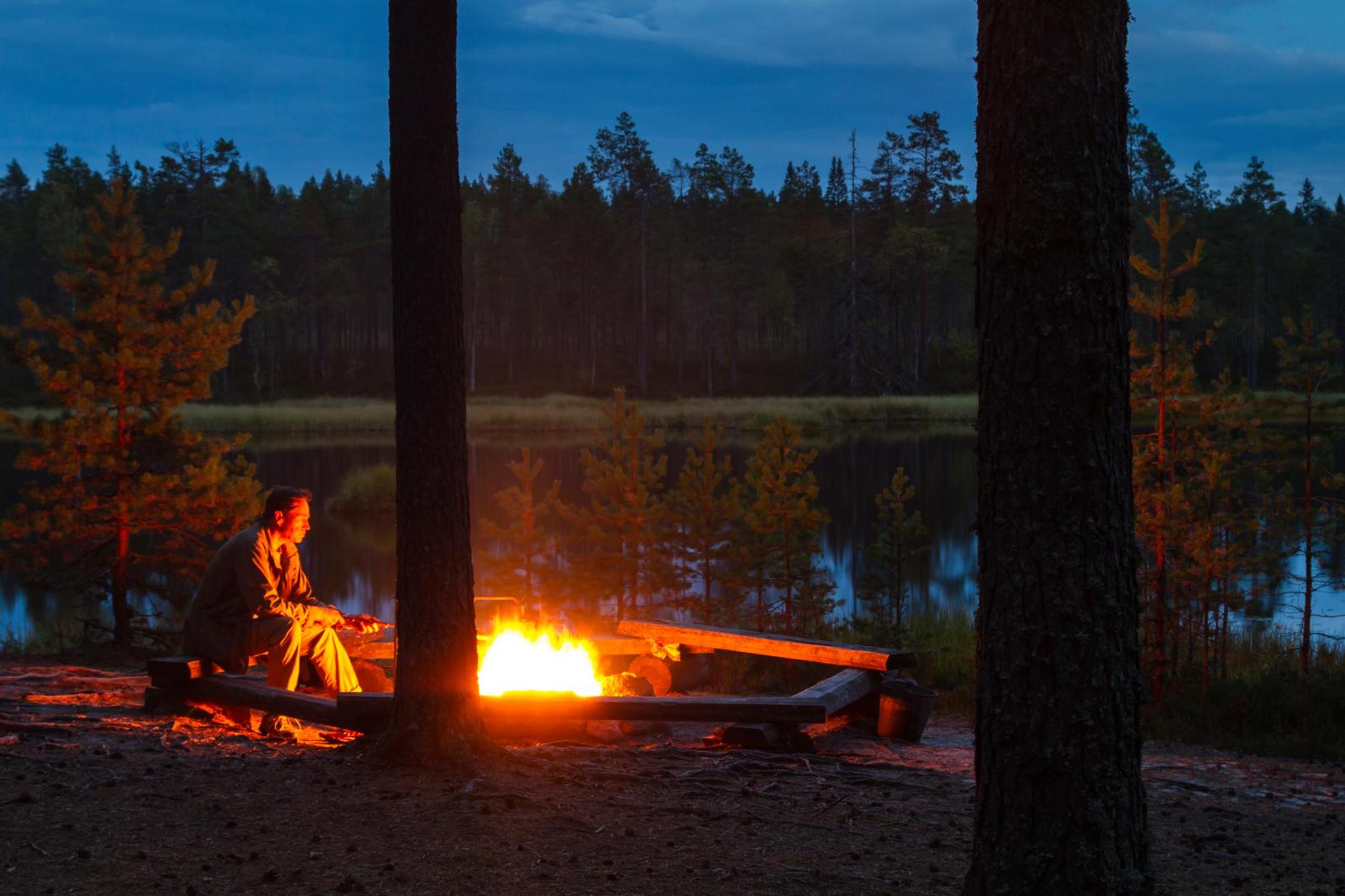 ihminen istuu tulipaikalla nuotion ääressä hämärässä illassa järven rannalla