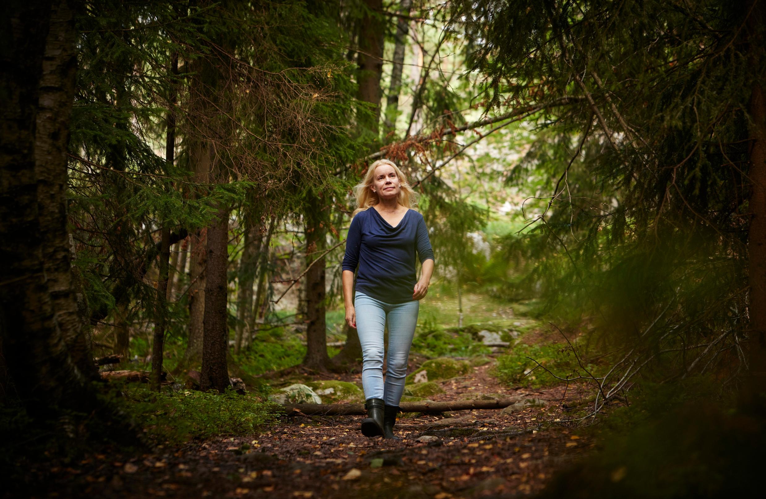 nainen kävelee pitkin metsäpolkua, jonka reunoilla kasvaa kuusia