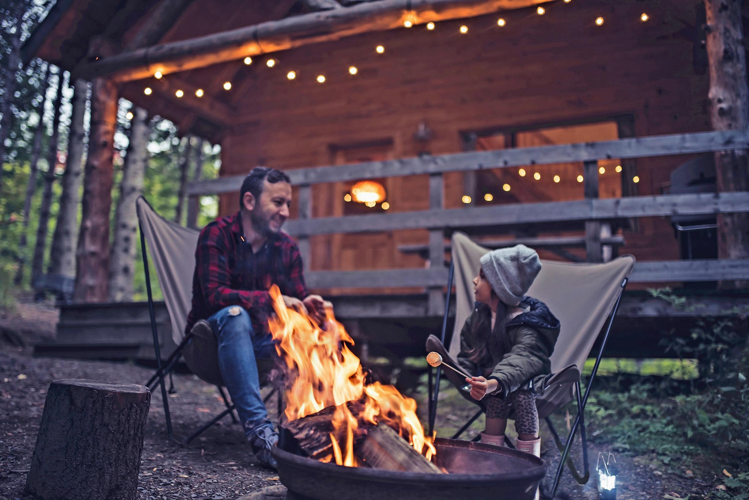 mies ja lapsi paistavat tikkupullaa nuotiolla mökin edessä hämärässä illassa