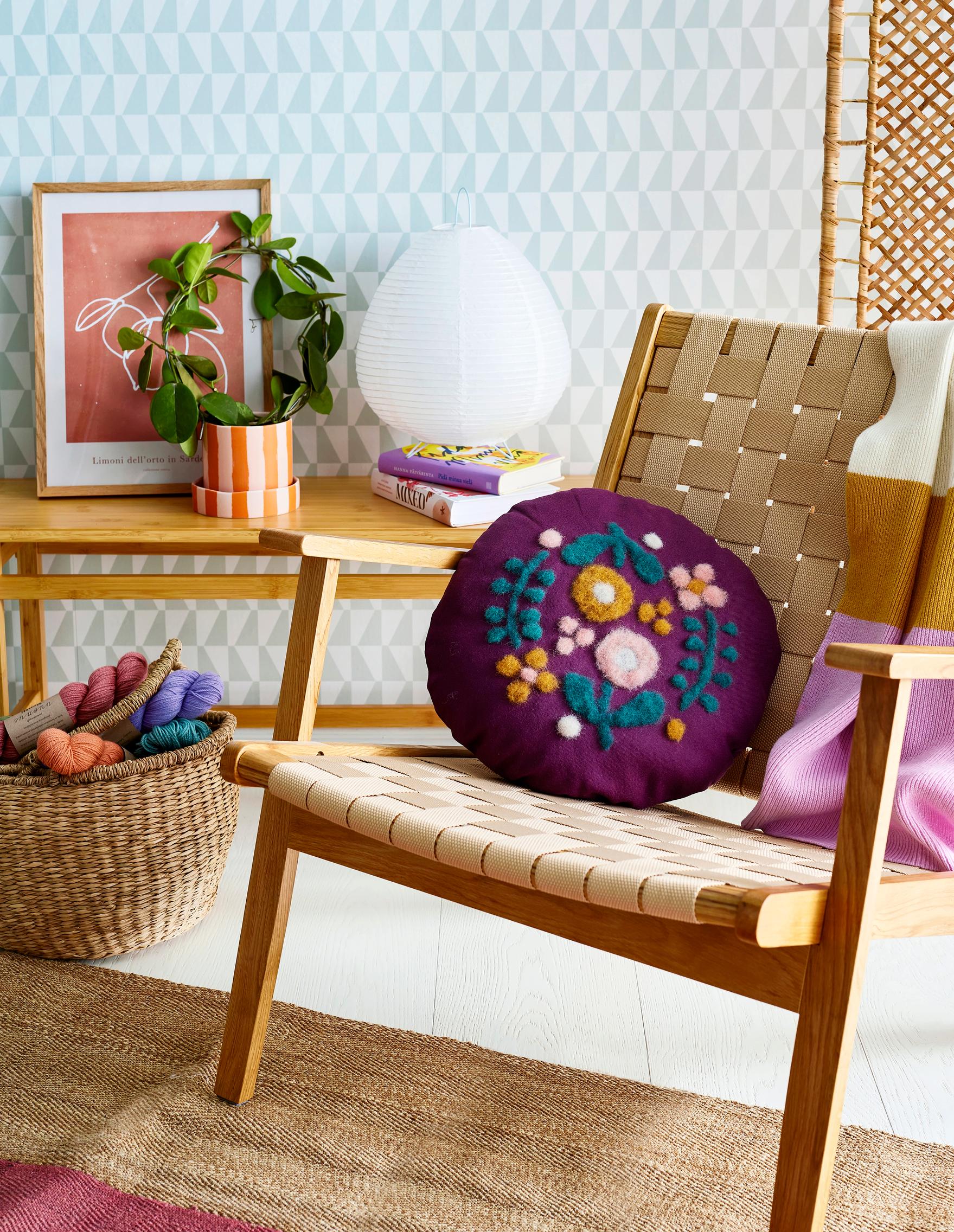 Violetti pyöreänmuotoinen tyyny, jossa on neulahuovutetut koristeet.