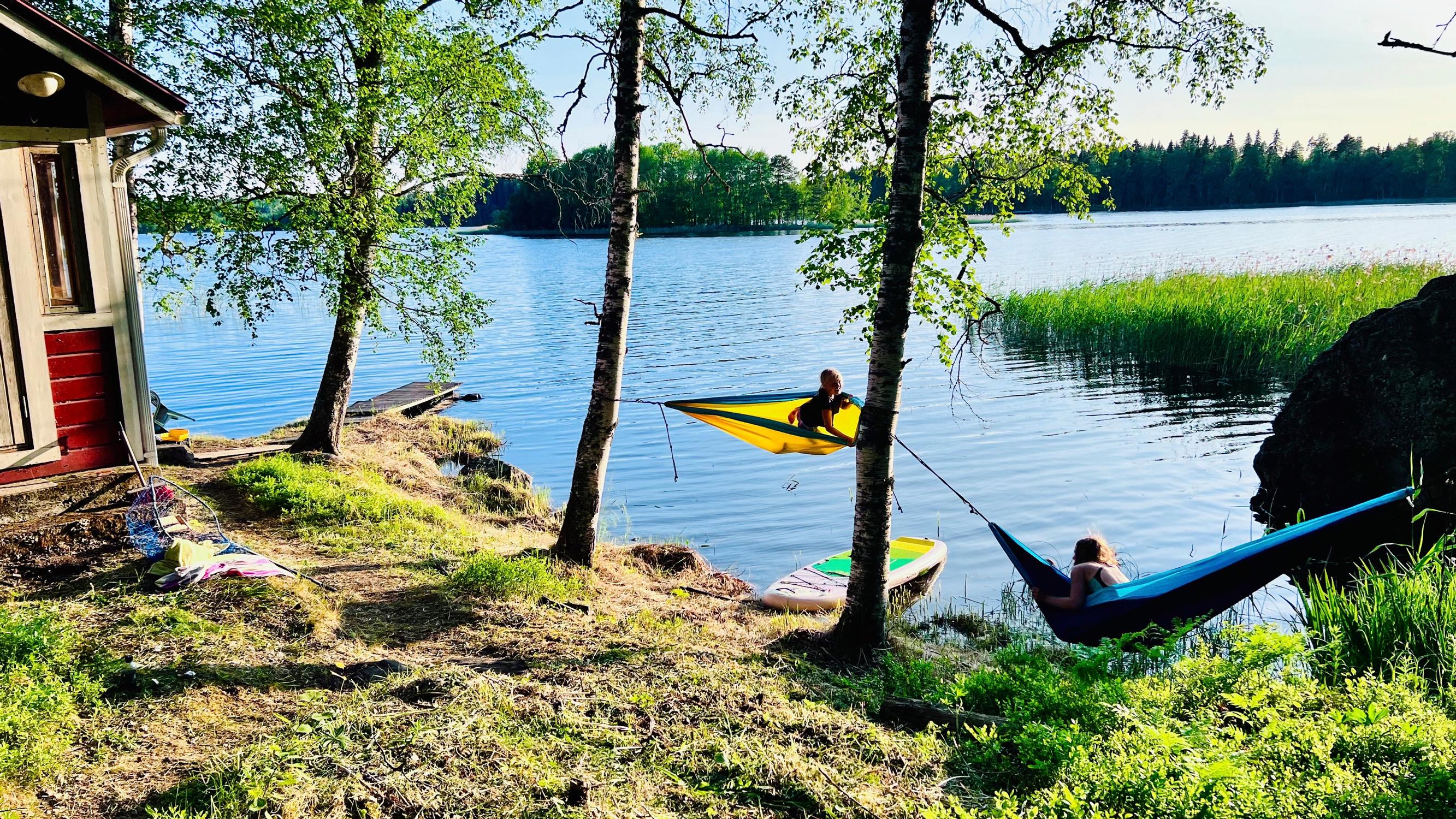 kaksi lasta riippumatoissa järven rannalla, punaisen rakennuksen kulma ja puita