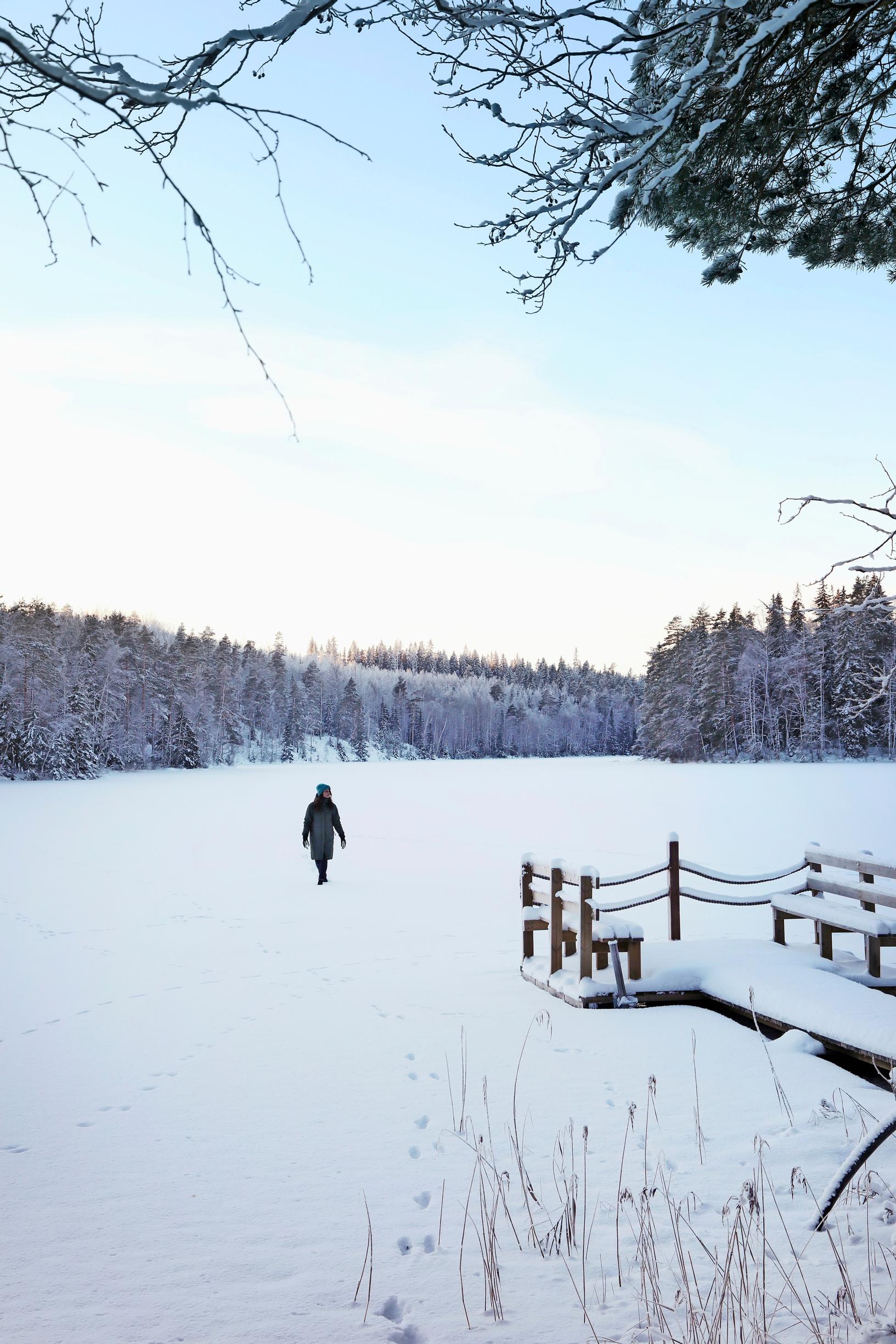 Ulla Parviainen kävelee keskellä lumista järvenselkää, etualalla laituri ja kaislikkoa, vastarannalla metsää