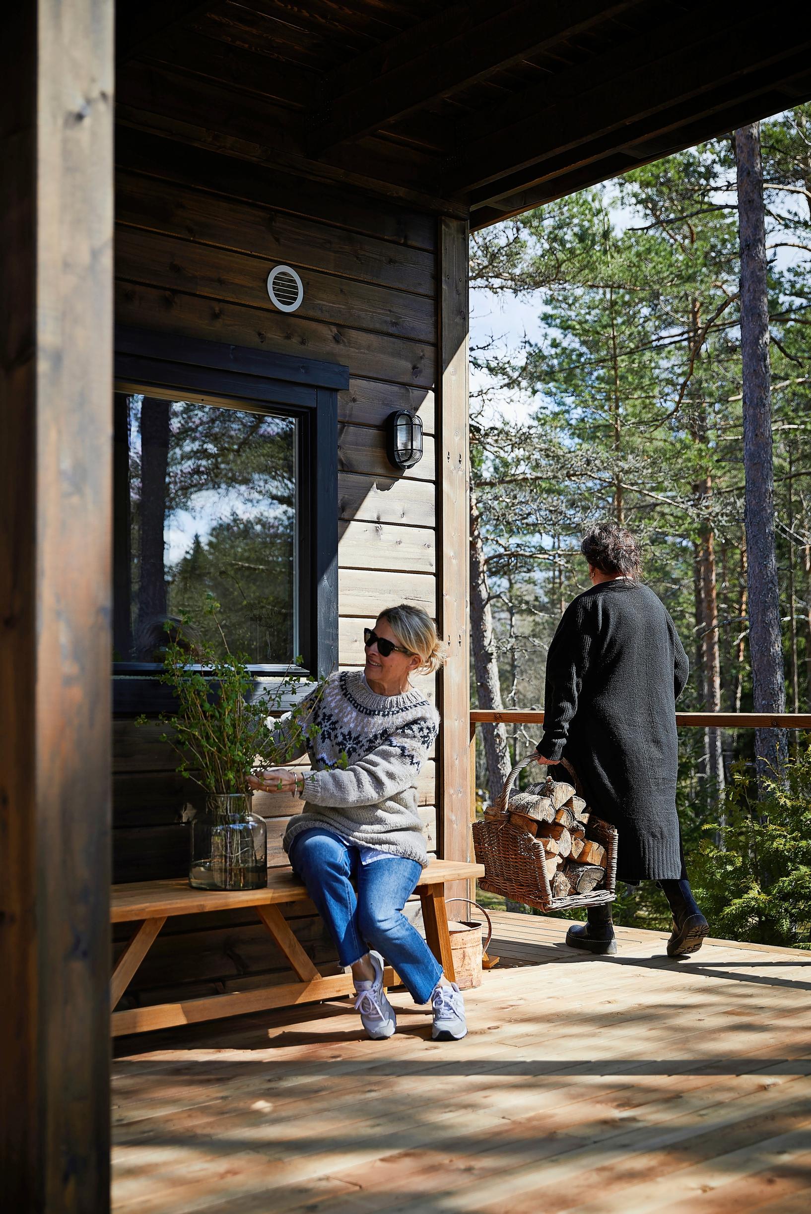 nainen istuu kasvin kanssa penkillä terassilla, toinen kantaa halkoja, taustalla mäntyjä