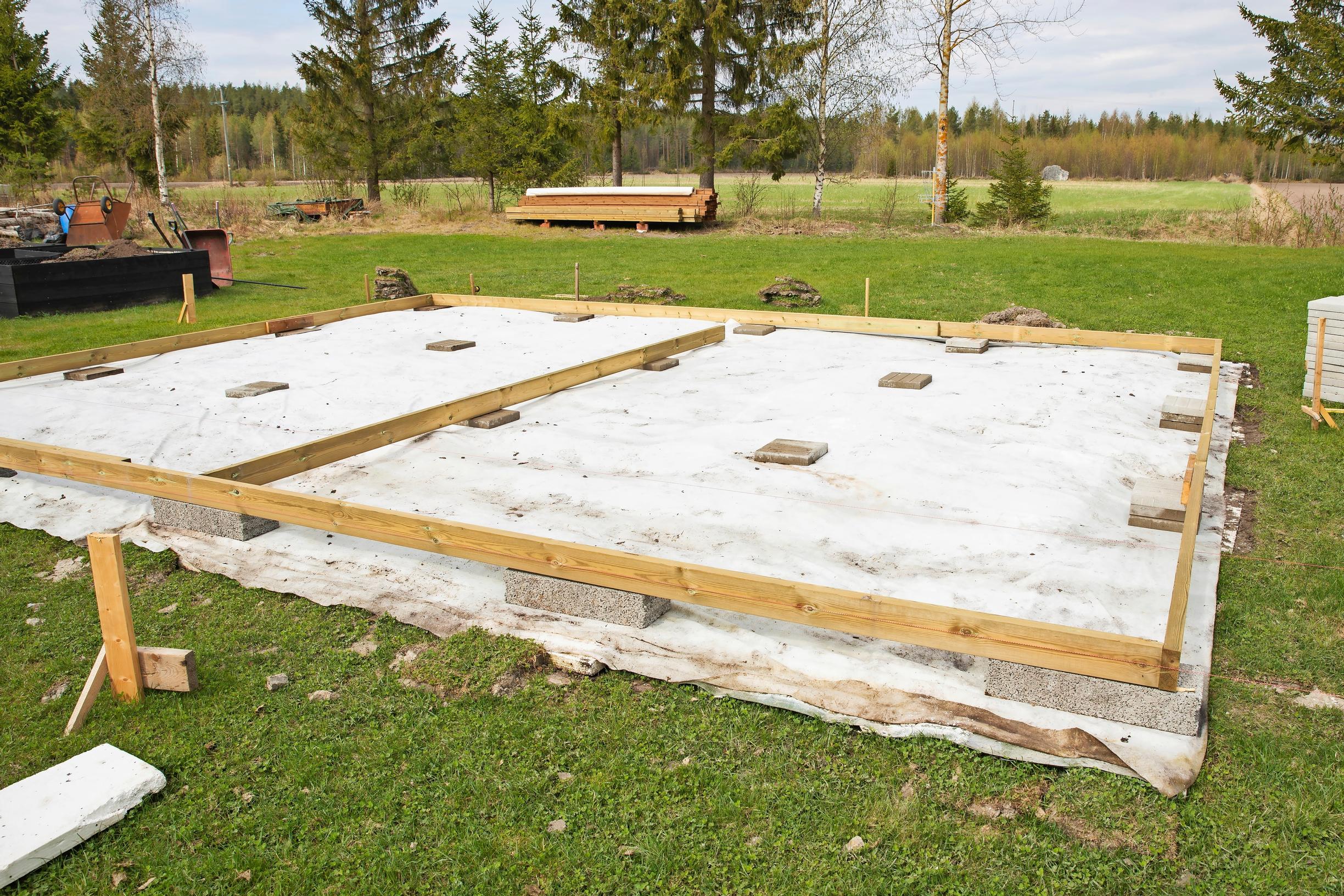 nurmikon keskellä valkoinen suojakangas, harkkoja ja puukehikko, taustalla nurmea ja metsää