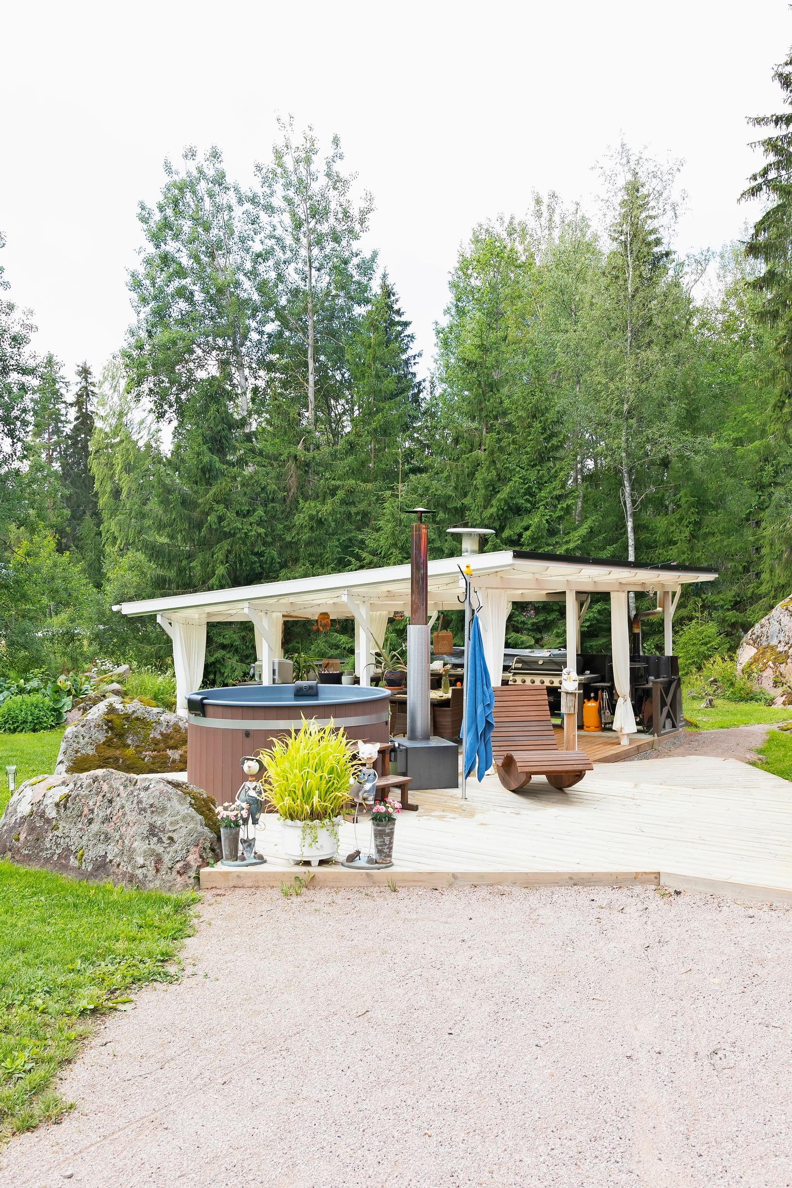 kesäkeittiö ja palju terassialueella, edessä sorapihaa, vieressä isoja kiviä, taustalla metsä