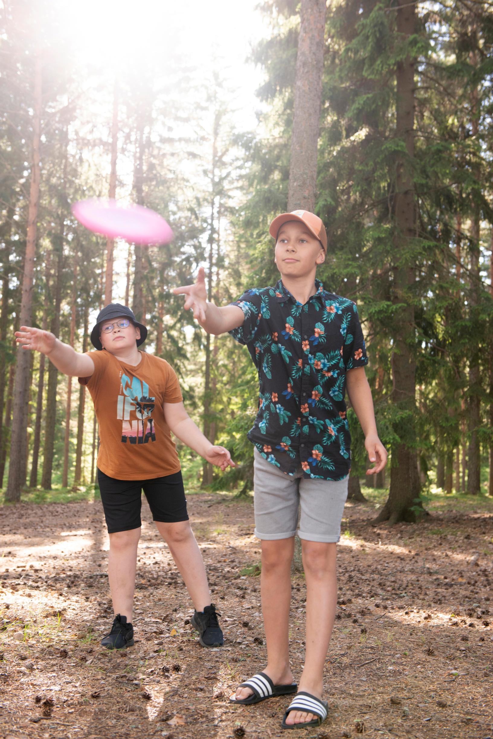 kaksi poikaa heittää frisbeetä mäntymetsä taustalla