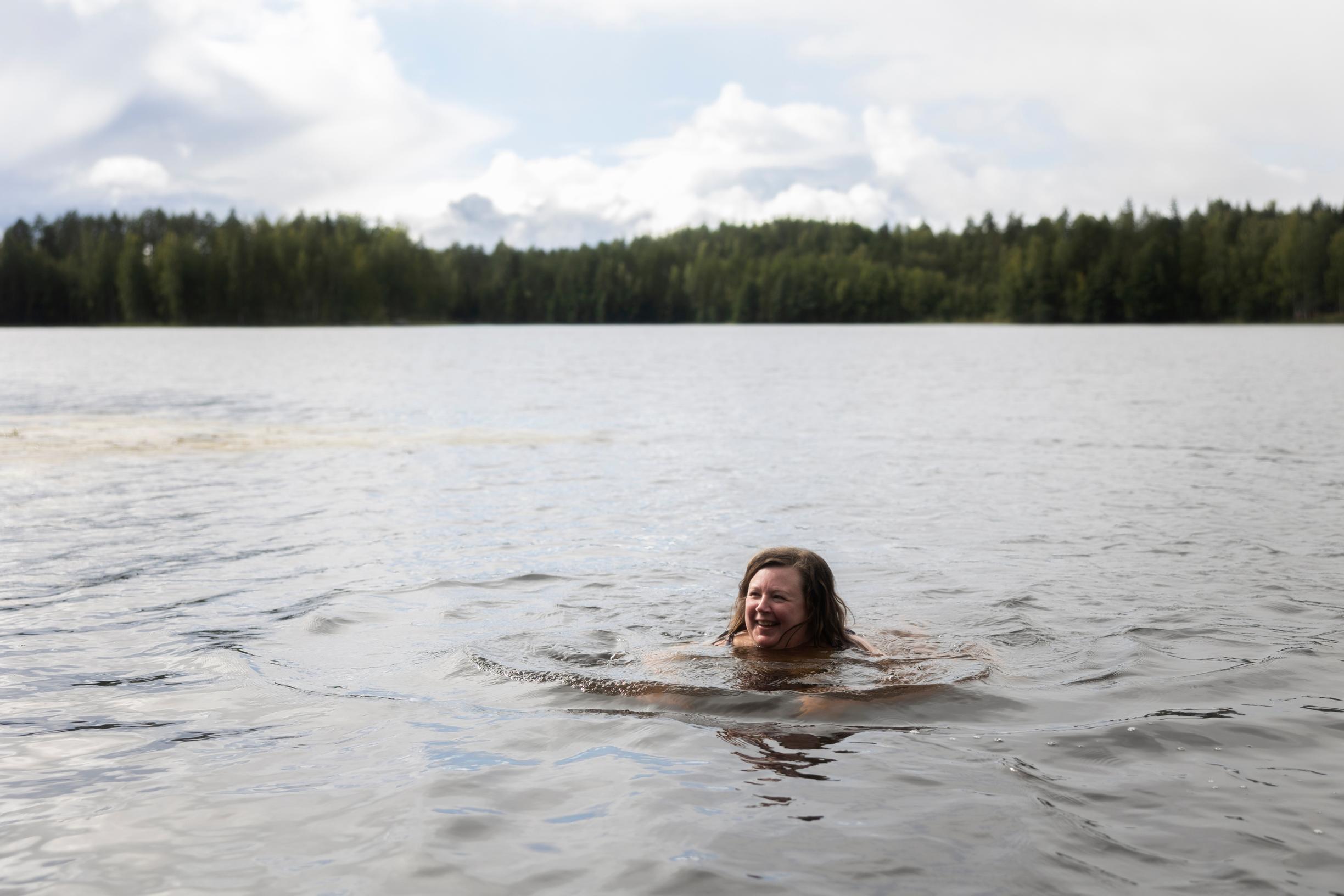 hymyilevä nainen ui järvessä, vastarannalla metsää