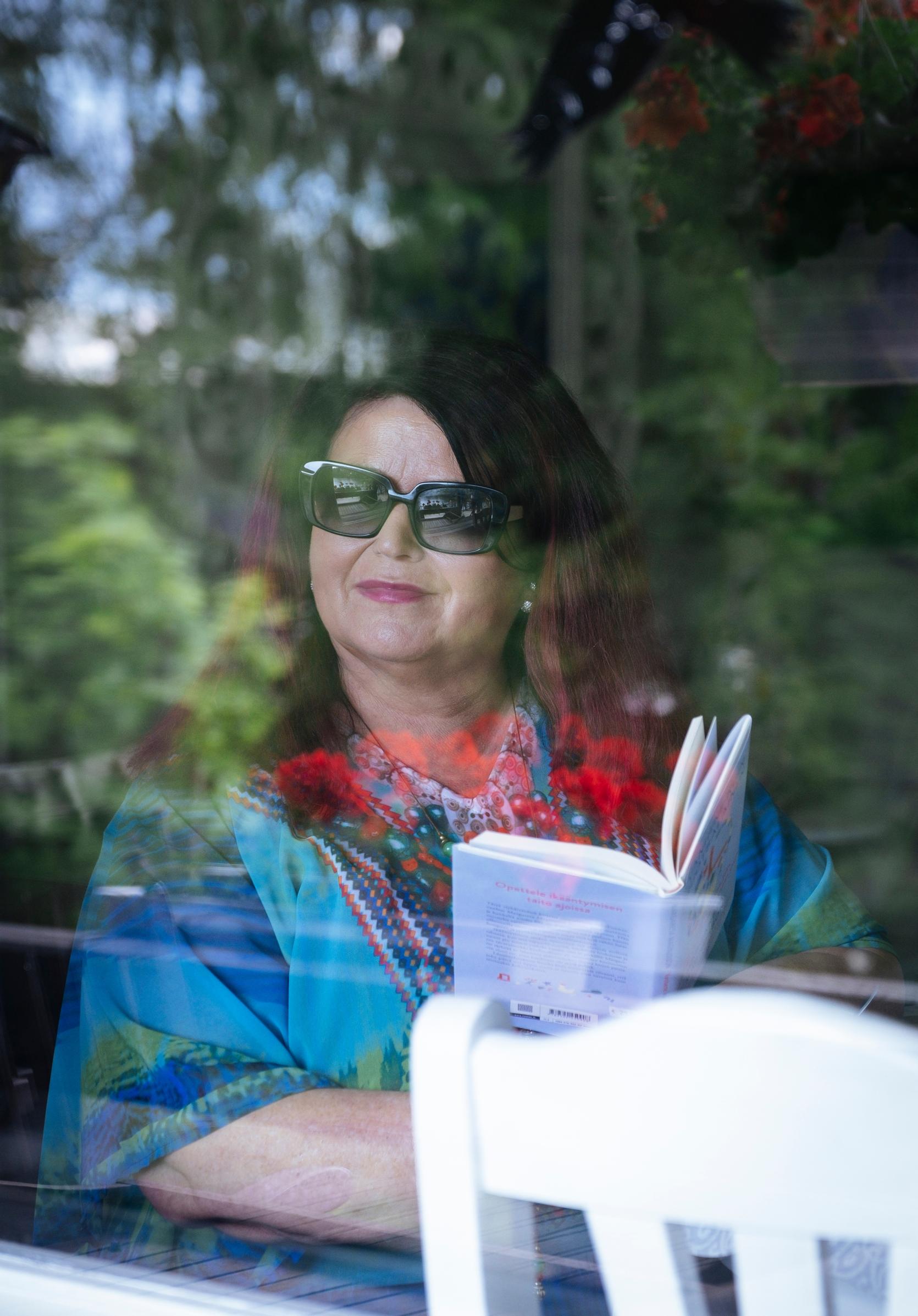 laulaja meiju suvas istuu ikkunan takana hymyillen kirja kädessä ja aurinkolasit päässä, edessä tuoli