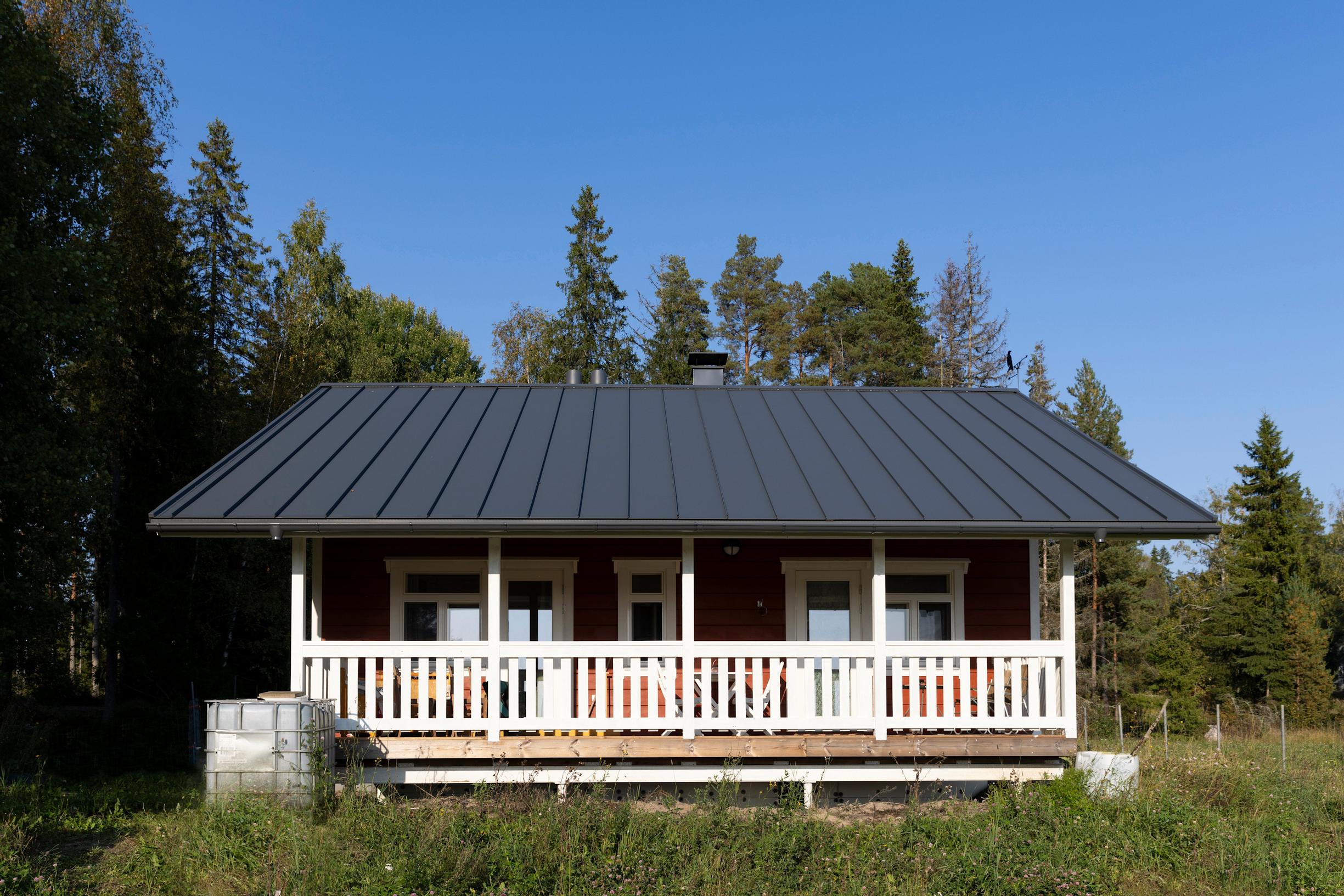 Harmaakattoinen talo pellon ja metsän reunassa, terassilla valkoiset puukaiteet.