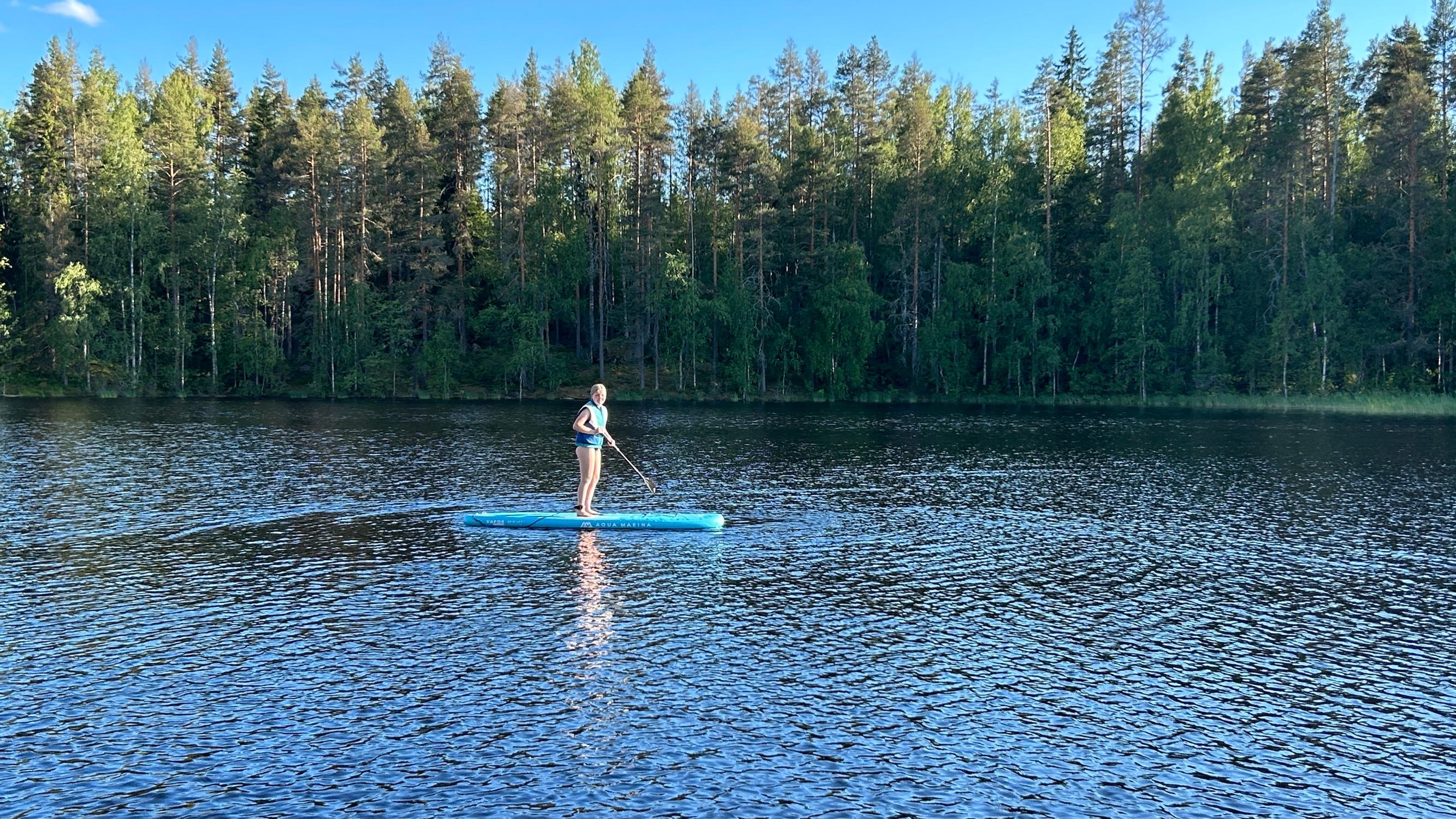 Alma Kaartinen meloo seisoen sup-laudalla järvellä, vastarannalla metsää