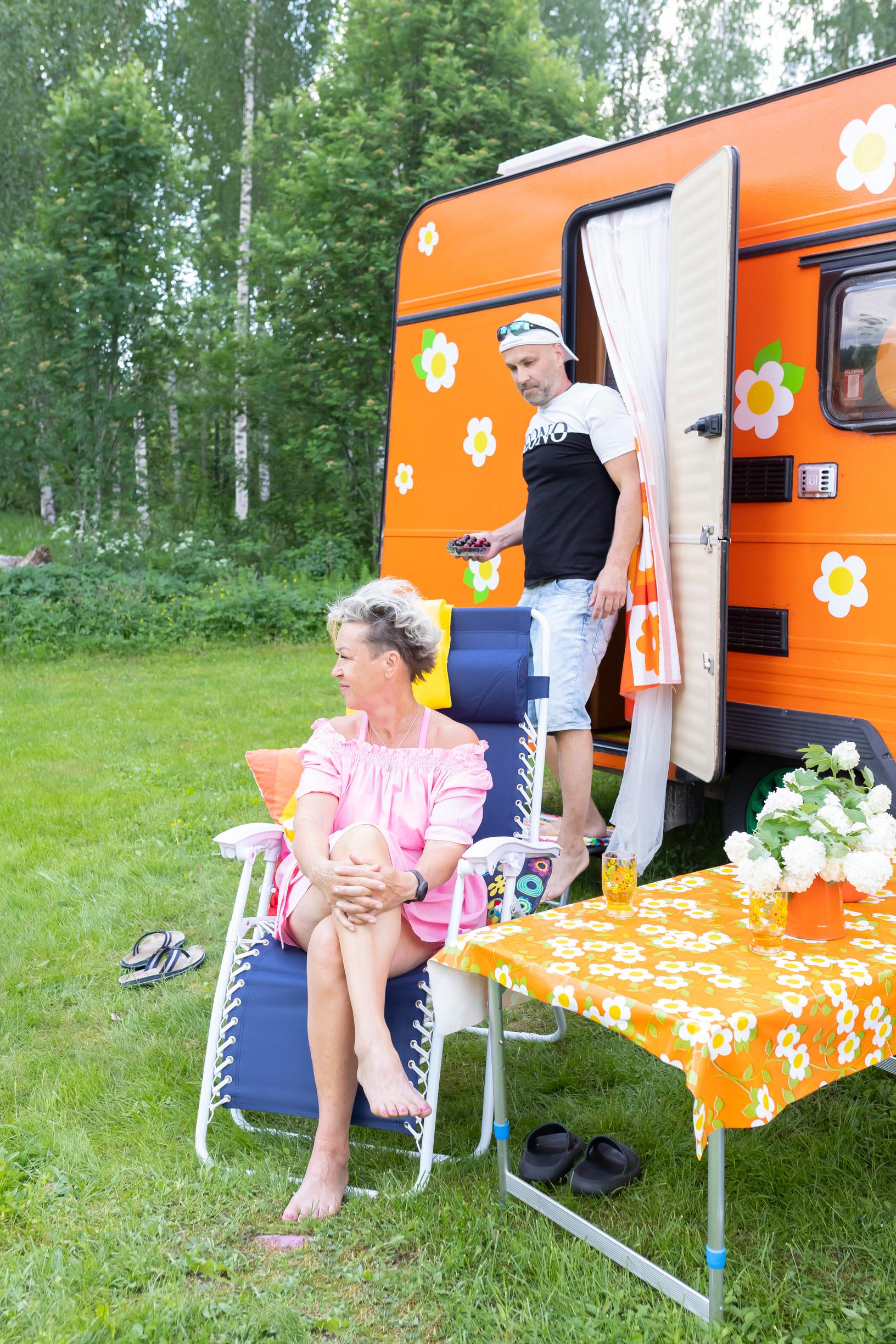 Kirsi Niiranen istuu puutarhatuolissa värikkään asuntovaunun edessä ja Jaakko Timonen tulee ulos vaunusta