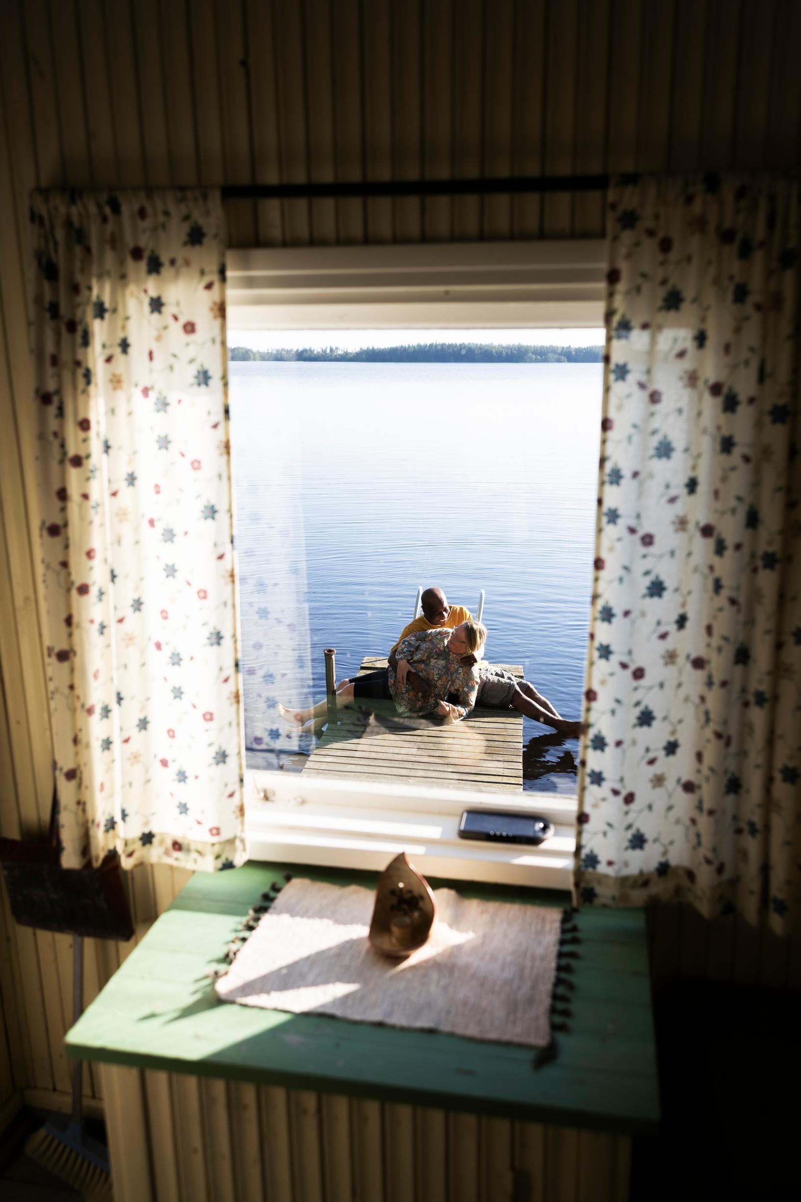 pariskunta näkyy istumassa laiturilla, näkymä ikkunasta, vaaleat kesäverhot