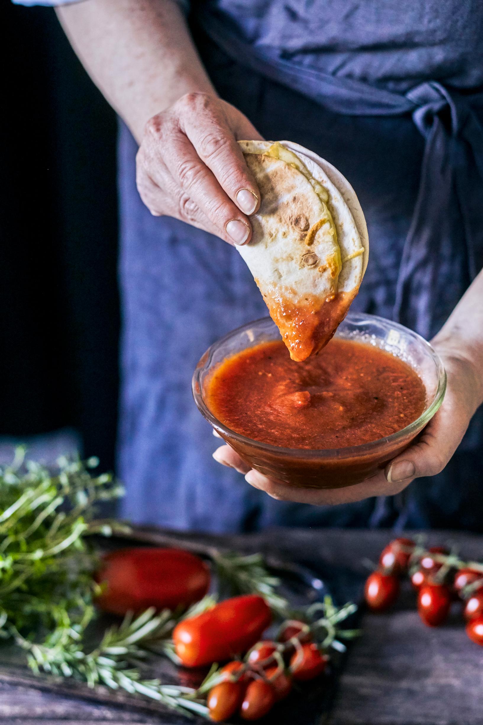 Pizzakastiketta kulhossa, käsi pitelee pizzapohjaa, joka dippaa kastiketta.