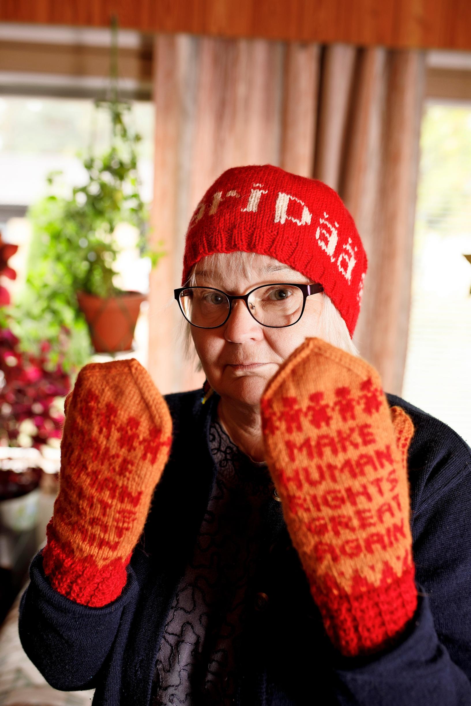 nainen katsoo kameraan ja näyttää lapasia, joissa lukee make human rights great again. hänellä on päässään punainen pipo, jossa lukee ääripää
