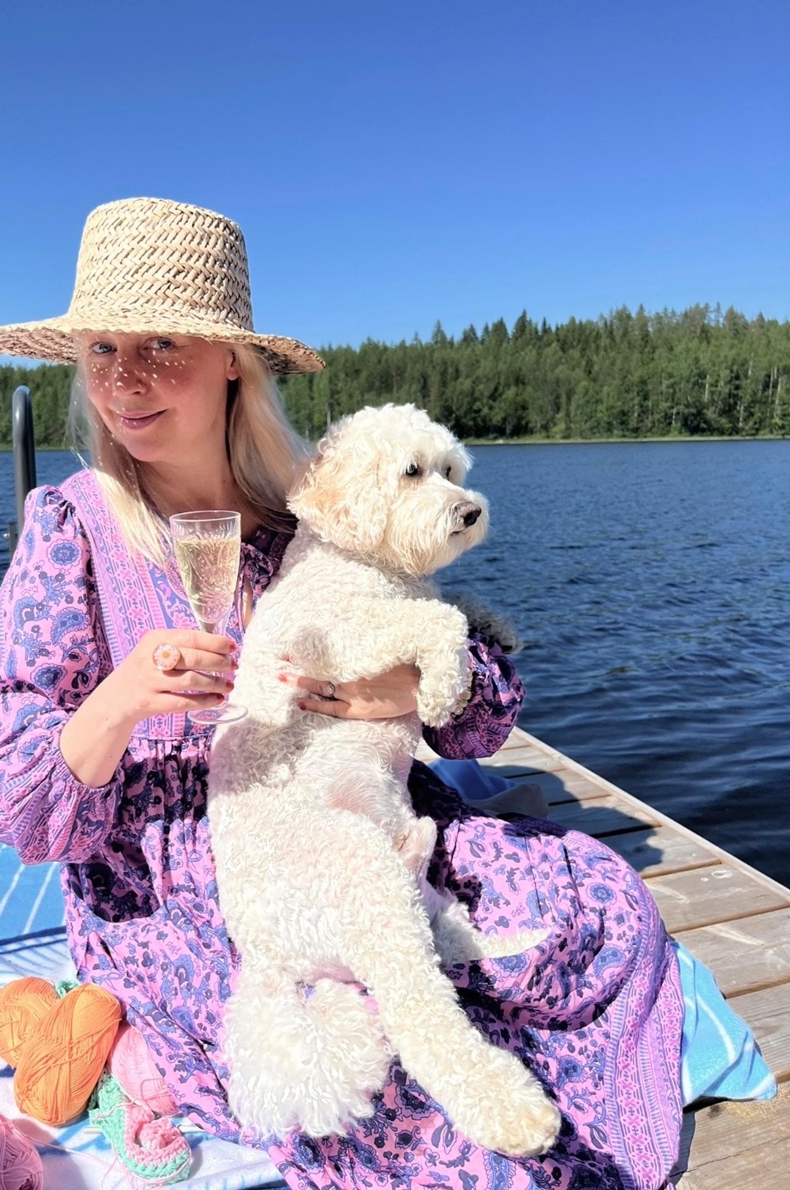 Näyttelijä Krisse Salminen istuu laiturilla piknikillä päässään olkihattu, kädessään kuohuviinilasi ja sylissään valkoinen koira