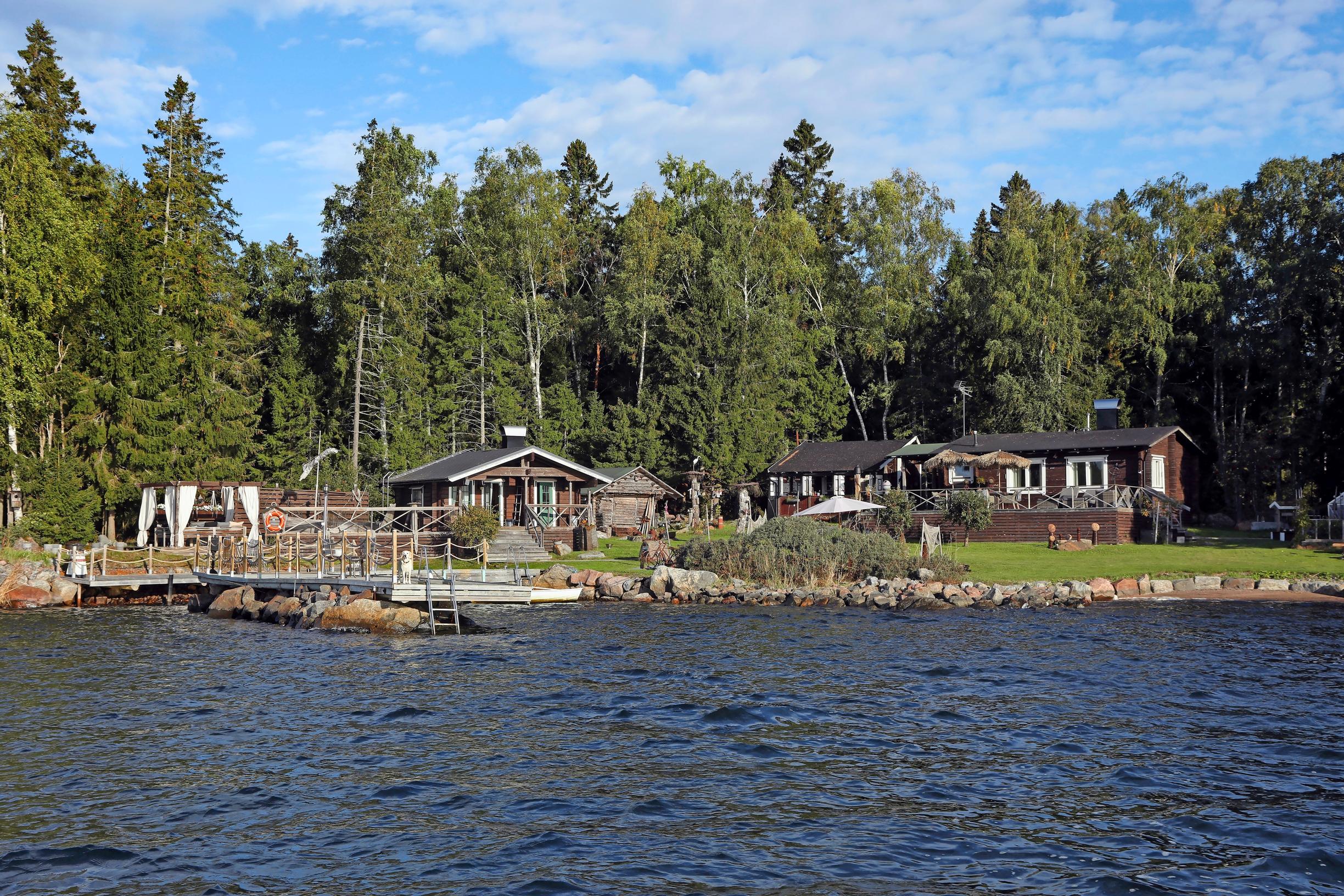 Ruskeita rakennuksia rannassa, laituri ja taustalla metsää.