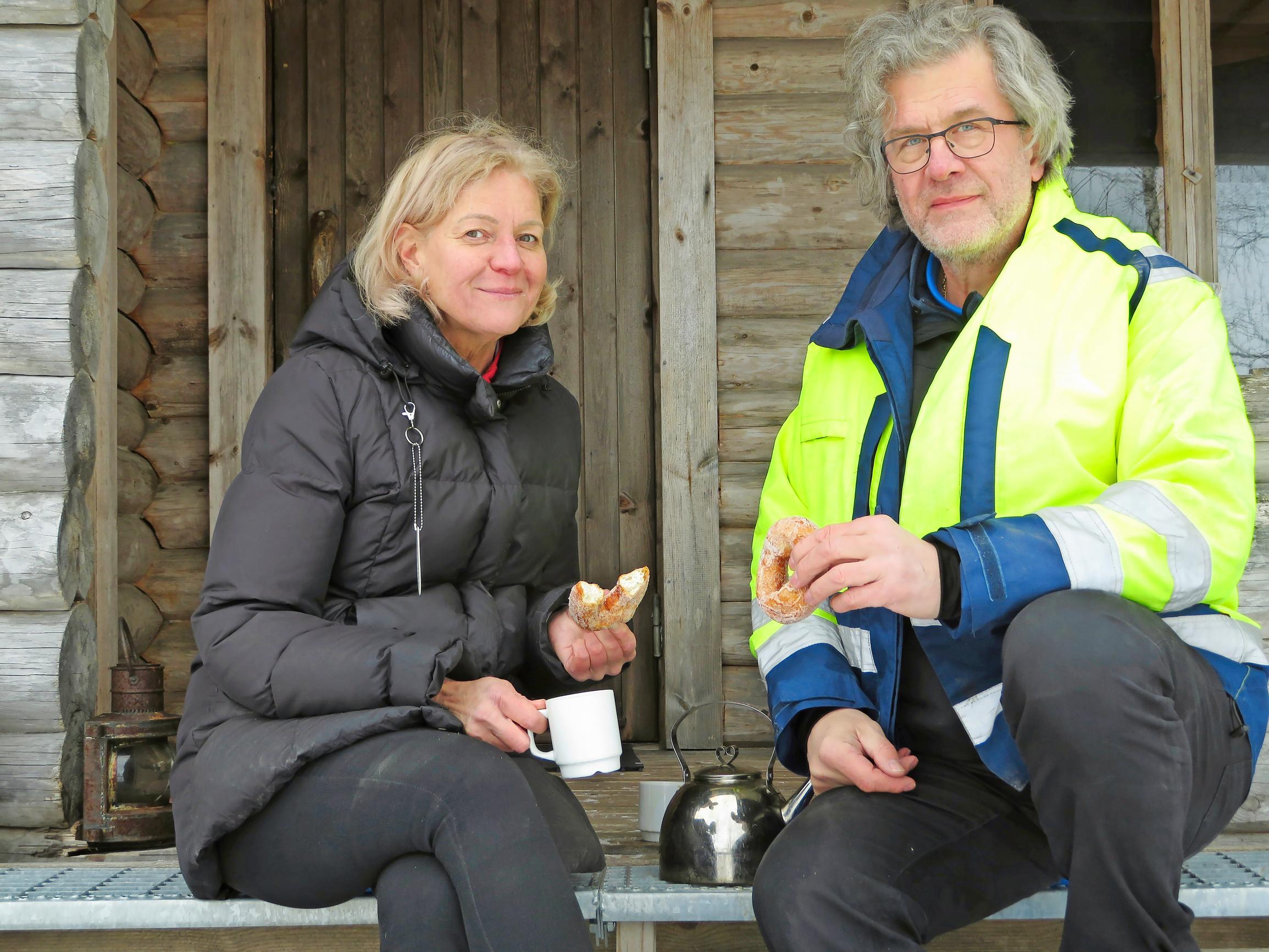 Cati Sundström ja Jyrki Hyytiäinen istuvat hirsimökin terassilla juomassa kahvia munkit kädessä