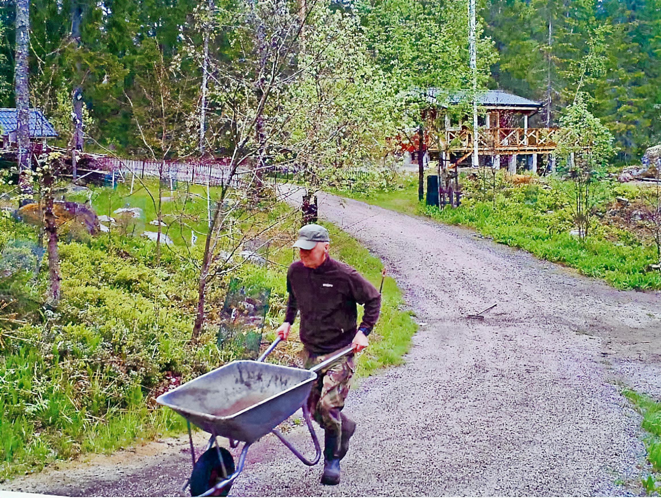 Näyttelijä Pirkka-Pekka Petelius työntää soratiellä kottikärryjä, taustalla  punainen mökki ja puutarhaa