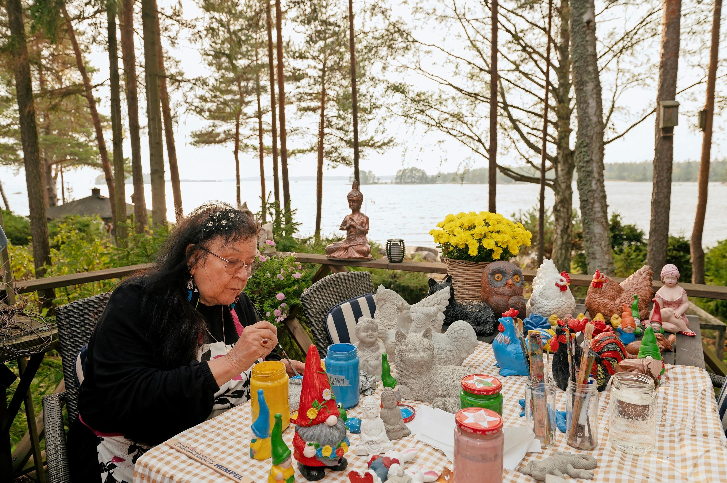 Arja Ögård maalaa betonityötään istuen terassin pöydän ääressä meren äärellä, pöytä täynnä esineitä