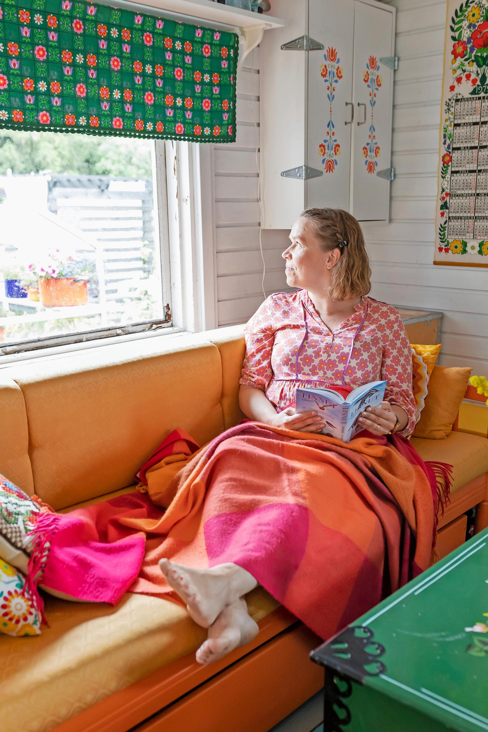 Leni Joutsjoki istuu kirja kädessä oranssilla sohvalla ja katsoo ulos