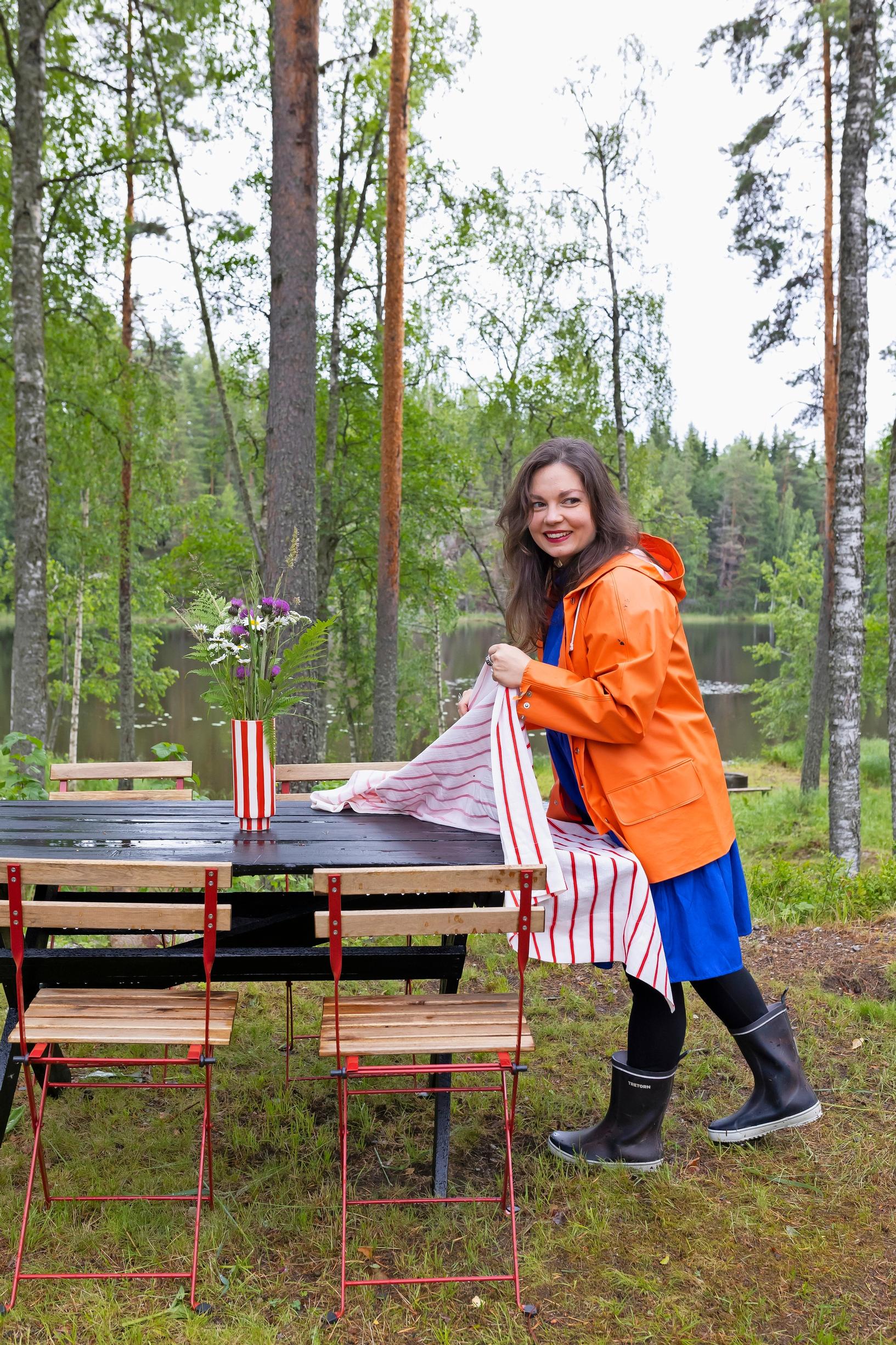 nainen laittaa ulkopöytään pöytäliinaa saappaat jalassa oranssissa sadetakissa, taustalla järvi
