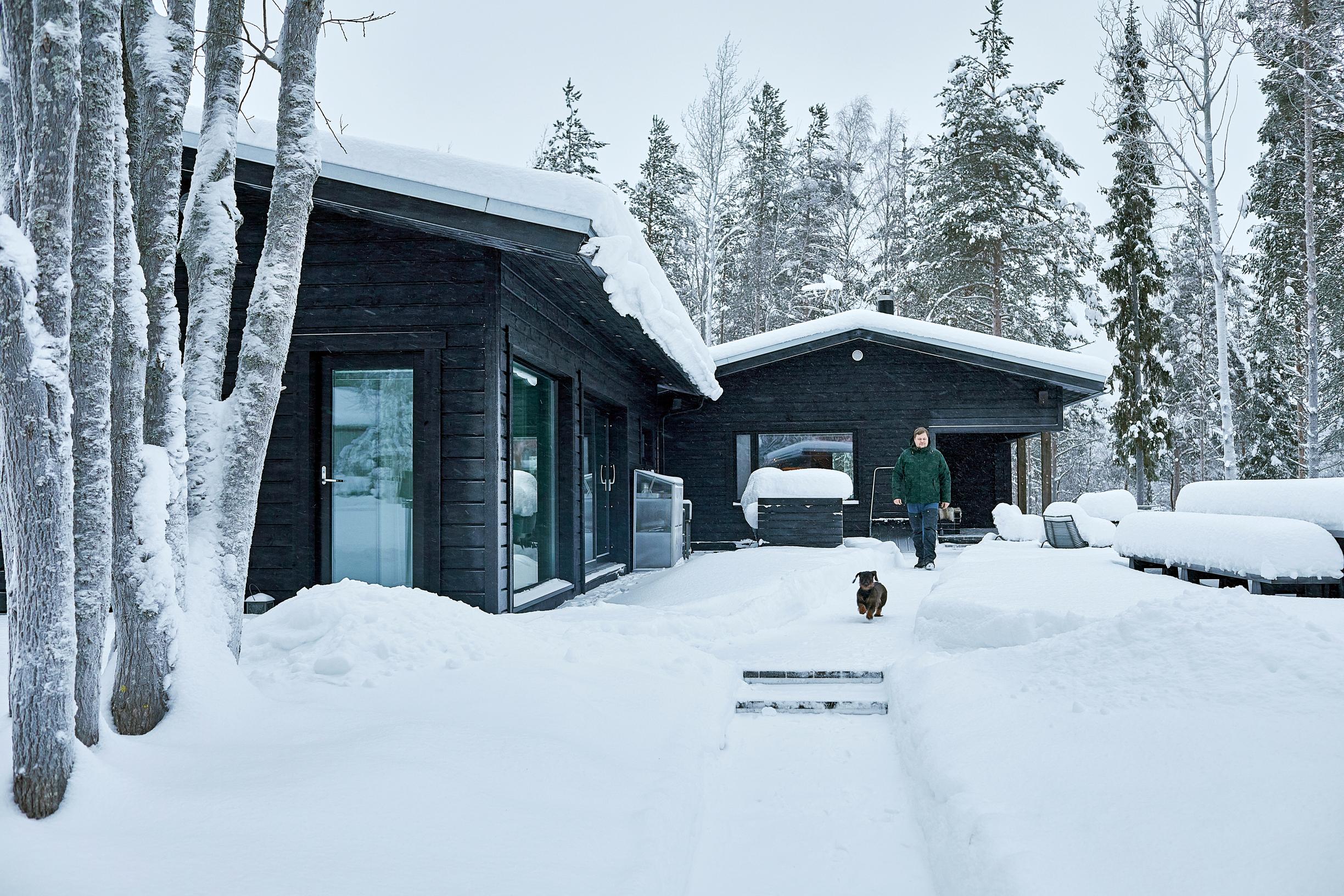 lunta mustien puurakennusten katolla ja pihalla, mies ja koira, taustalla metsää