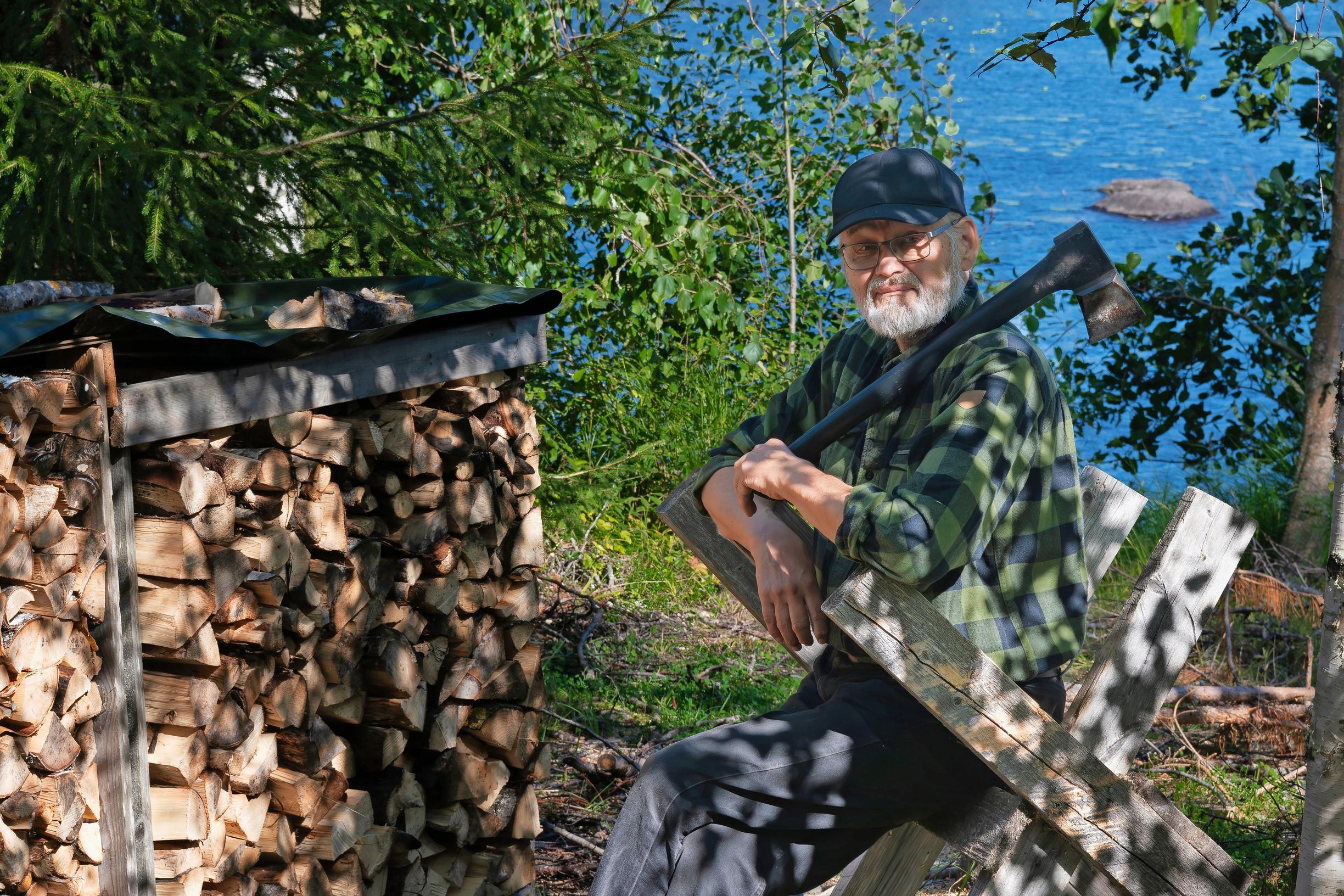 Pekka Vaaramo istuu sahapukilla kirves olalla, vieressä halkopino ja taustalla järvi