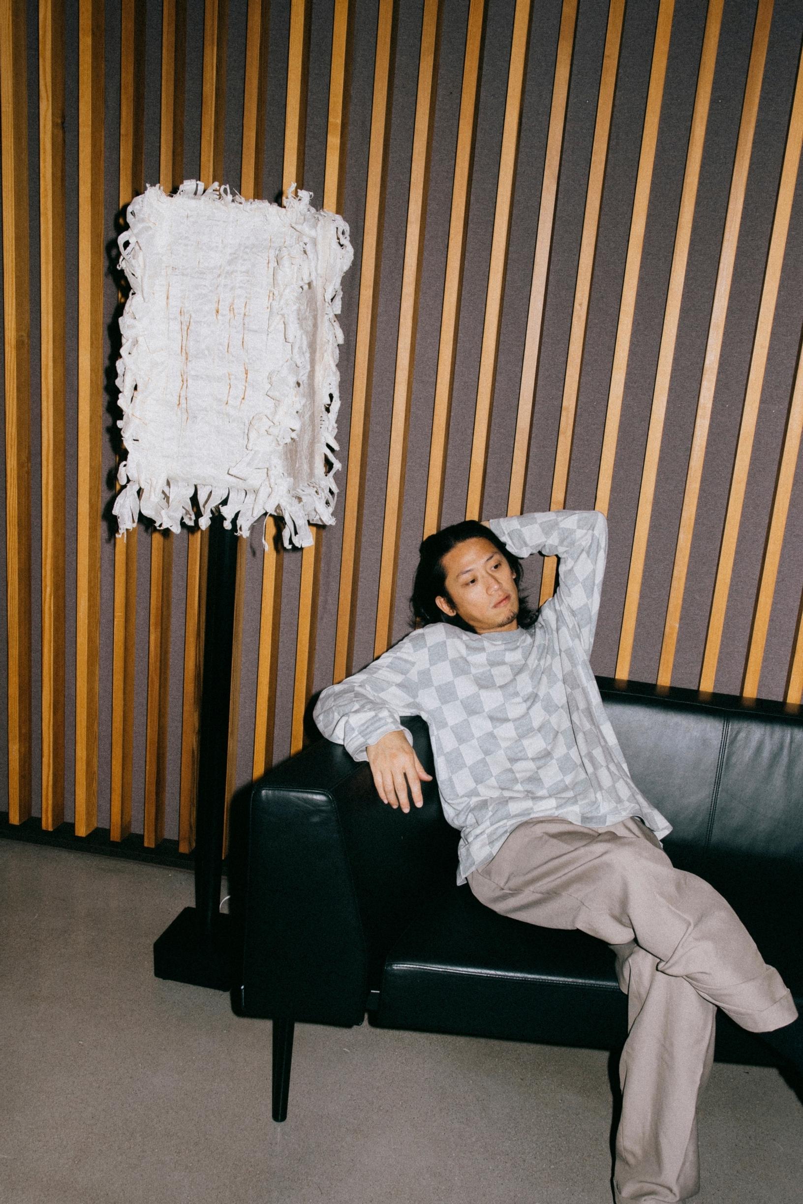 mies istuu mustalla sohvalla rennosti käsi pään takana, taustalla puusäleikköä, vieressä mustavalkoinen jännä jalkalamppu