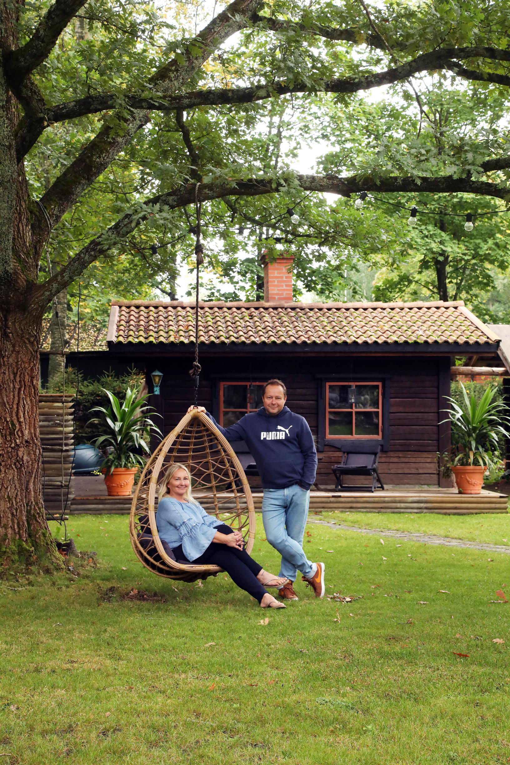Katja istuu puusta roikkuvassa korituolissa, Kimmo seisoo vieressä. Taustalla on puinen saunarakennus.