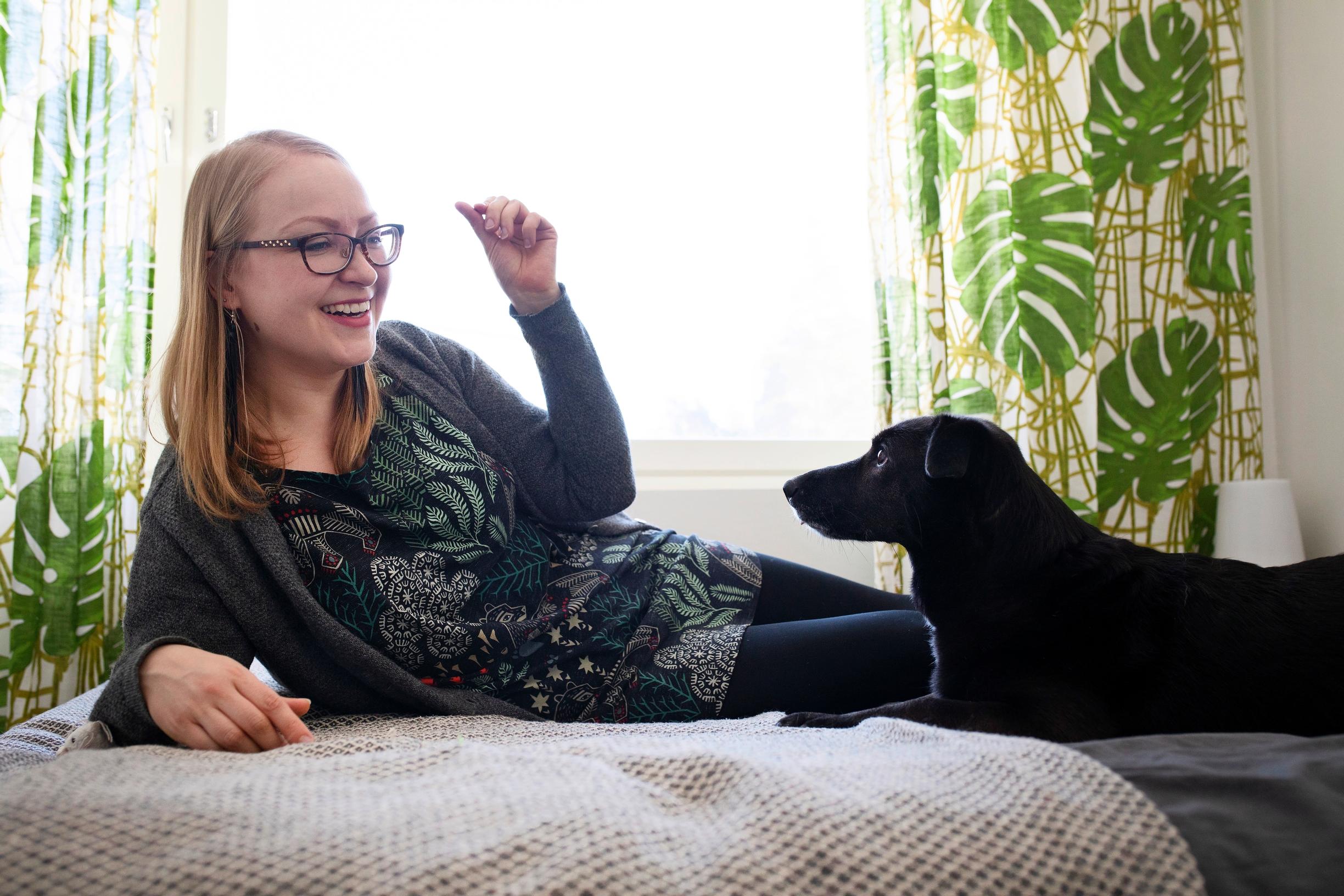 Nainen ja musta koira sängyllä, taustalla peikonlehtiverho