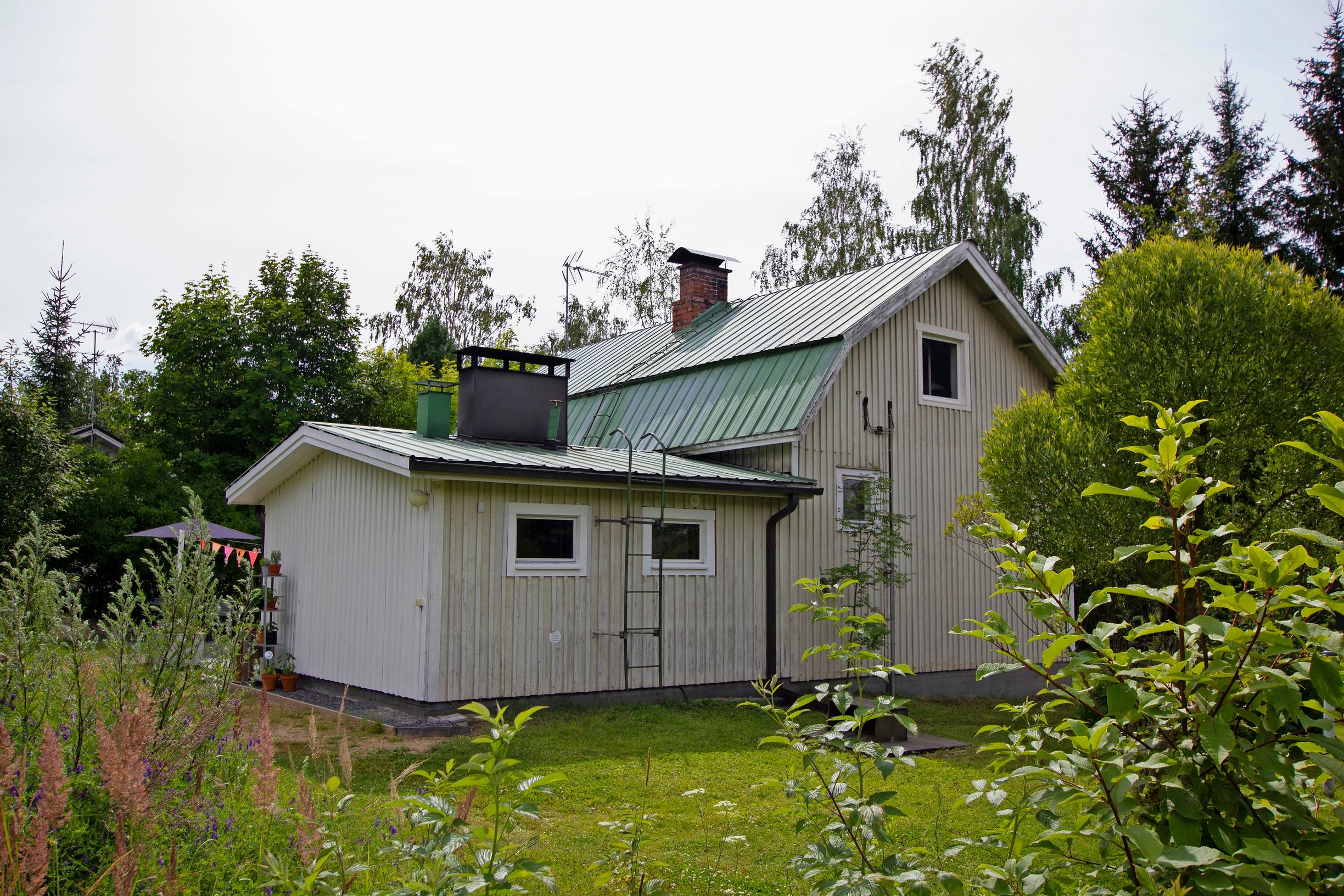 vaalea puutalo, jossa vihreä mansardikatto ja matalampi siipiosa, pihanurmi, kukkapenkkejä ja taustalla metsä