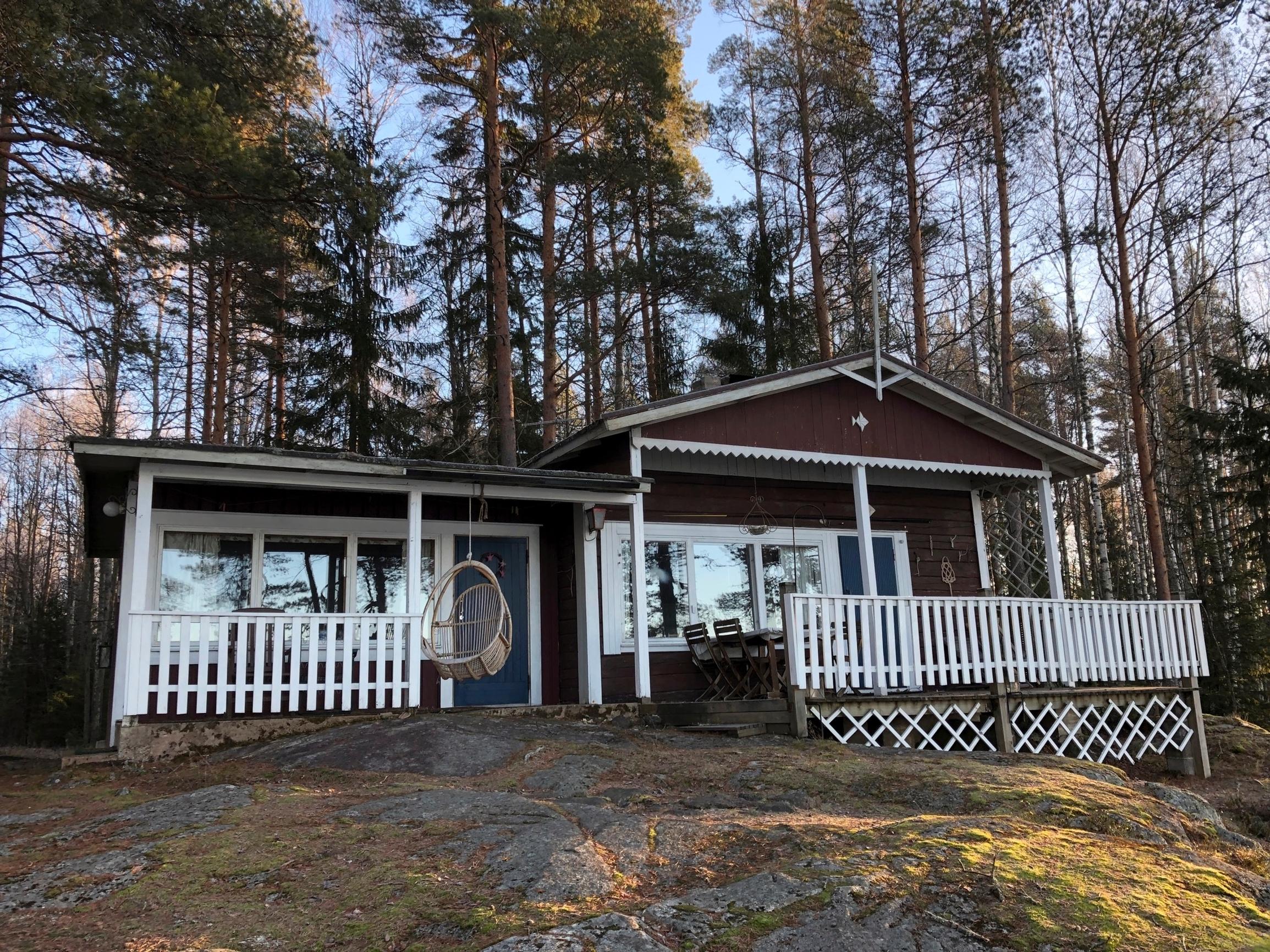vanha punaruskea mökki, jossa valkoiset terassit ja ikkunanpielet, etualalla kallio ja taustalla metsää