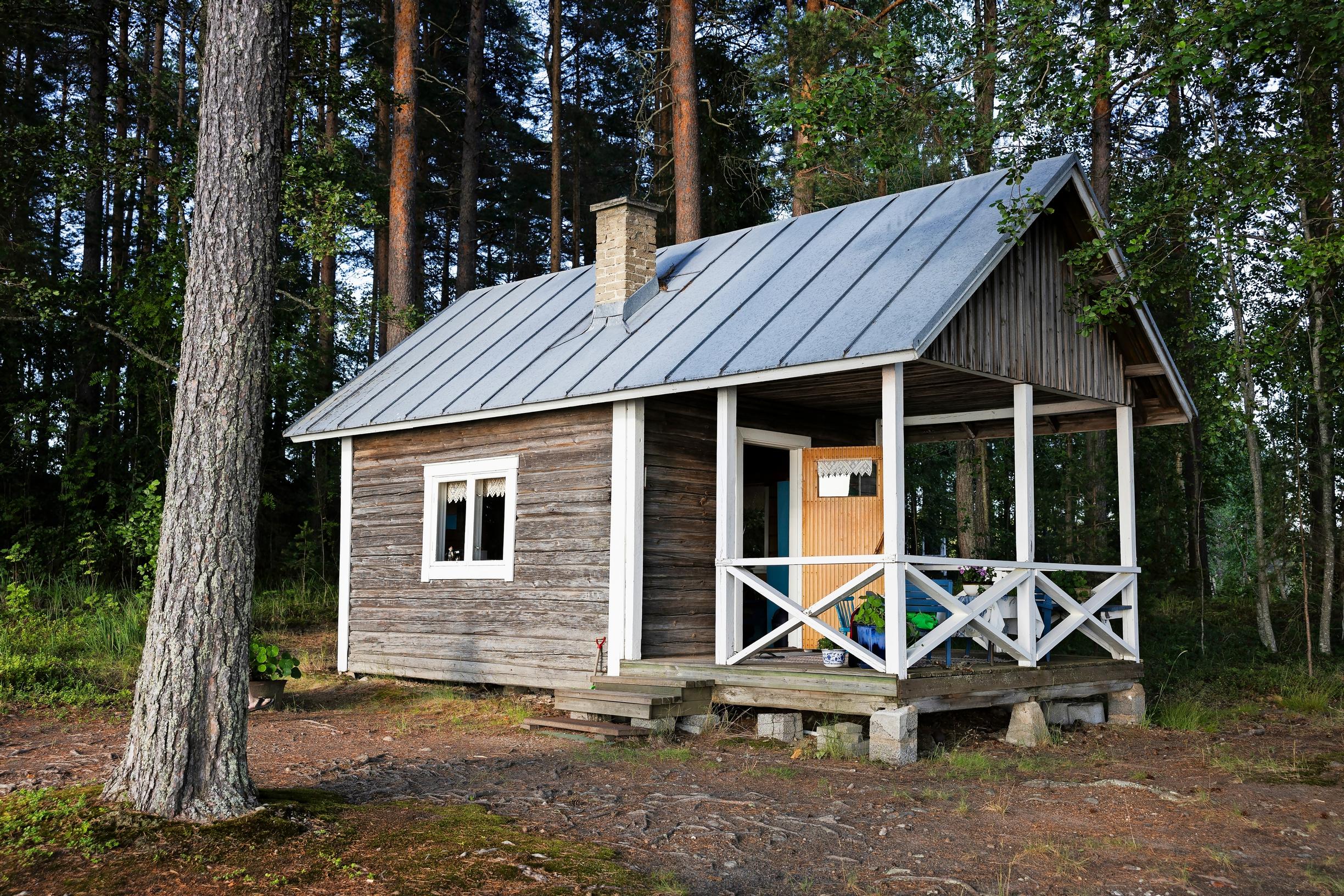 harmaa vanha hirsitalo, jossa valkoinen kuisti, taustalla metsää