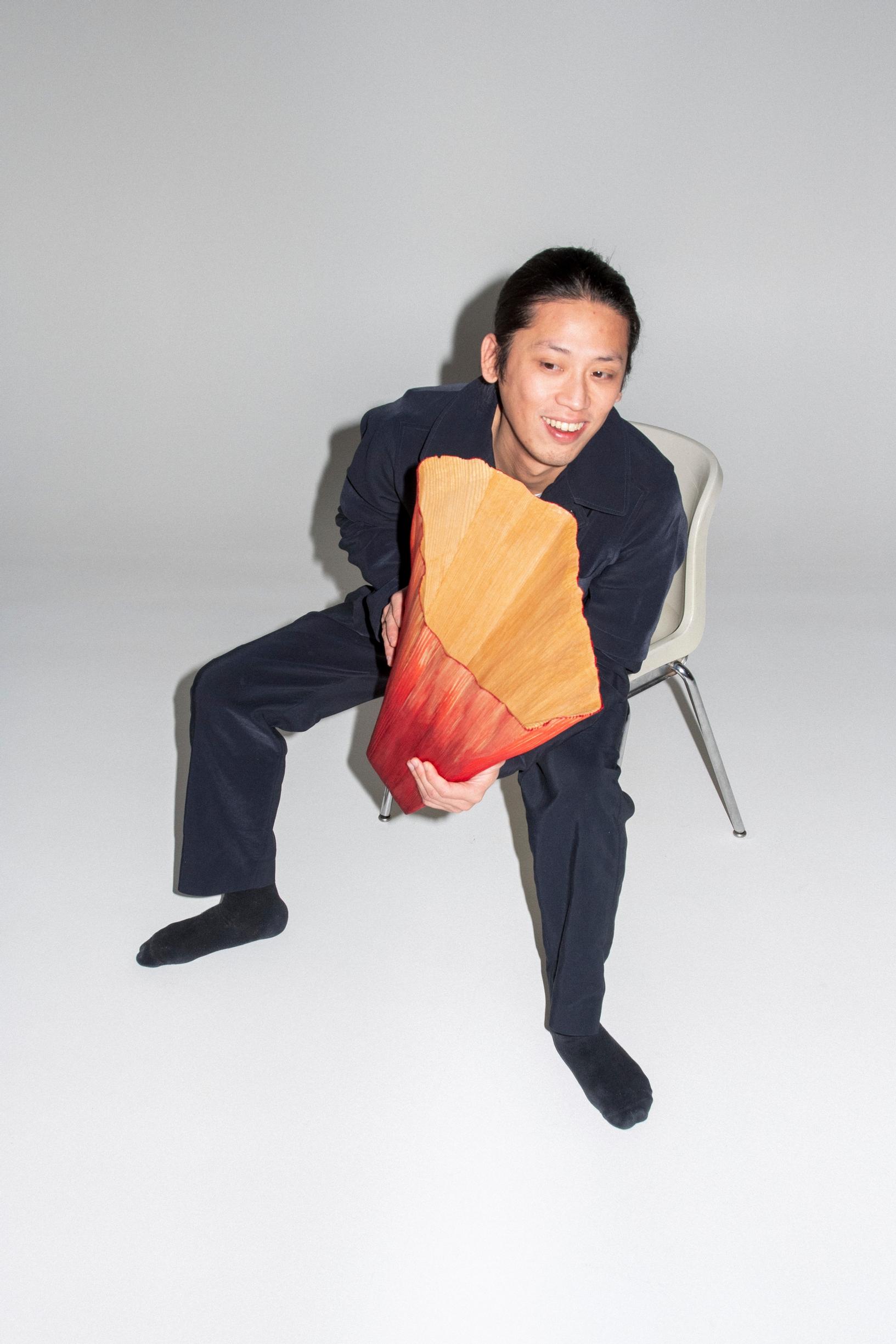 hymyilevä mustiin pukeutunut aasialaismies istuu tuolilla sylissään värikäs puuesine