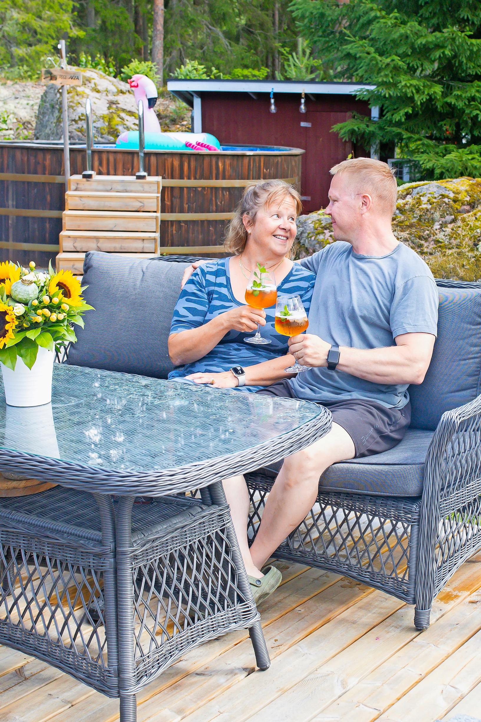 Päivi ja Petri Laamanen istuvat drinkit kädessä toisiaan katsoen mökin terassin puutarharyhmässä