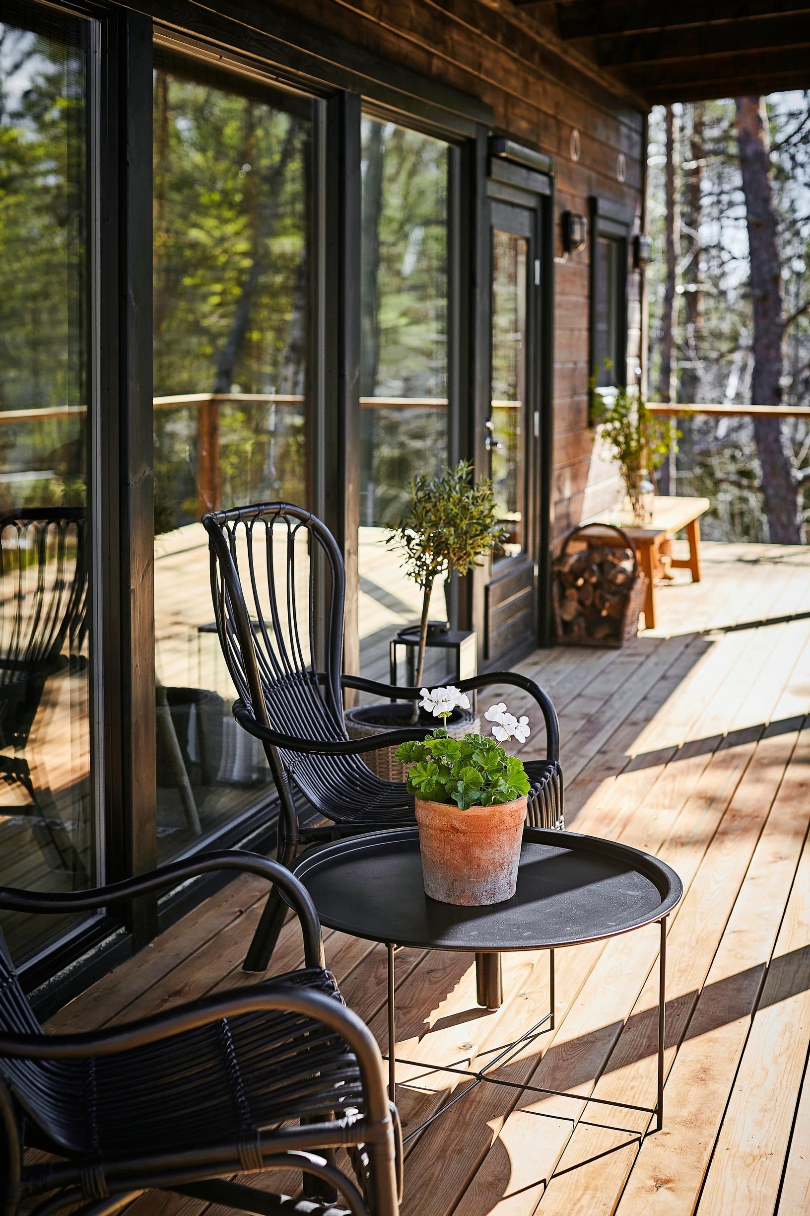 mustat rottinkituoli ja musta pyöreä pöytä mökin puisella parvekkeella, ruukkukukka, taustalla penkki