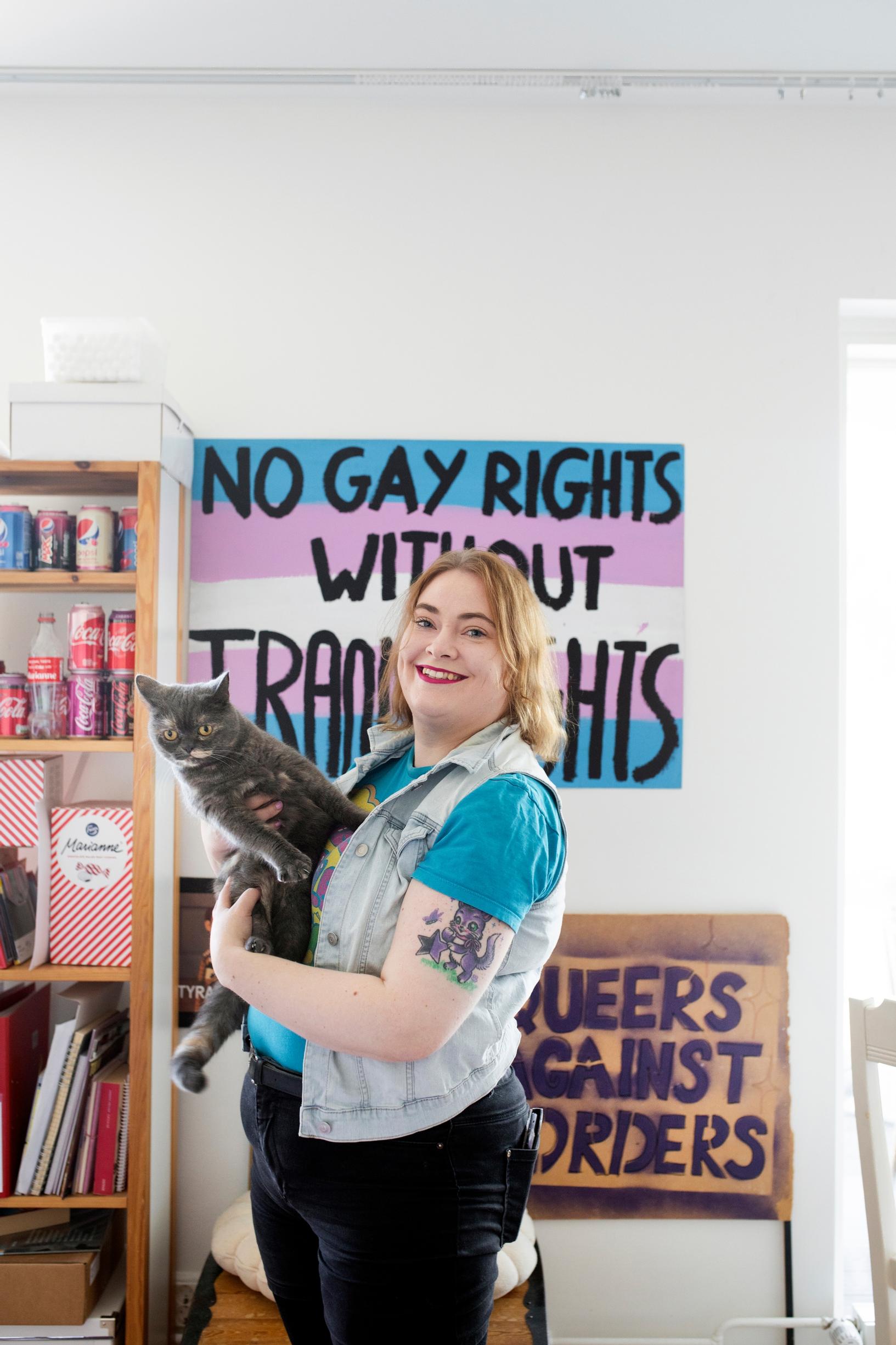 nainen pitää kissaa sylissään. taustalla olevissa julisteissa lukee no gay right without trans rights ja queers against orders