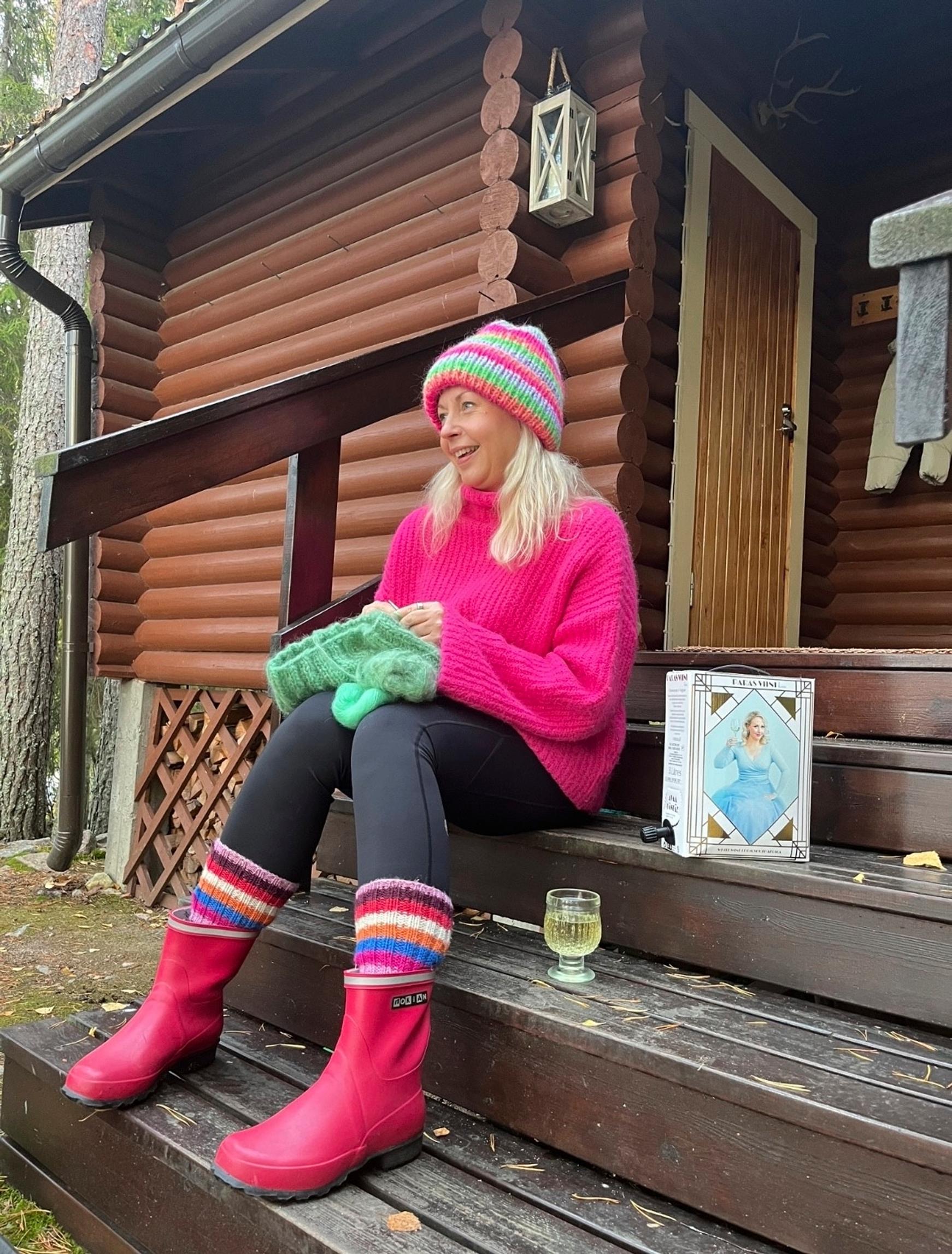 Näyttelijä Krisse Salminen istuu pinkeissä saappaissa ja pinkissä villapaidassa pipo päässä hirsimökin portailla, vieressä viinilasi ja -tonkka