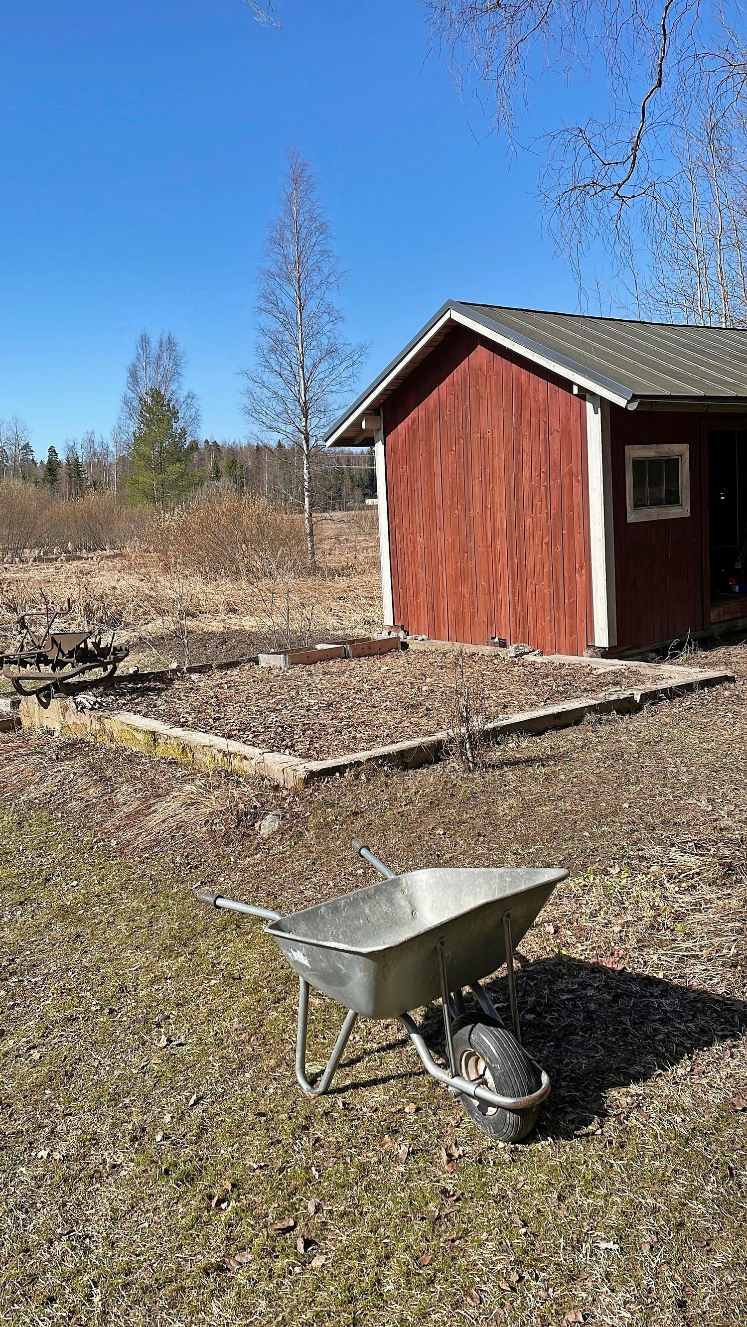 punainen vanha piharakennus, maassa kehikko sen vieressä ja kottikärryt