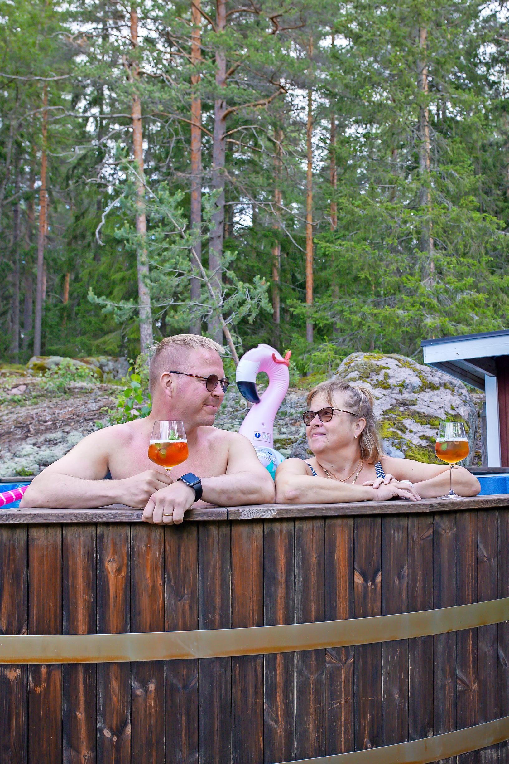 Petri ja Päivi Laamanen katsovat toisiaan uima-altaassa drinkit kädessä, taustalla flamingo-uimalelu ja metsää