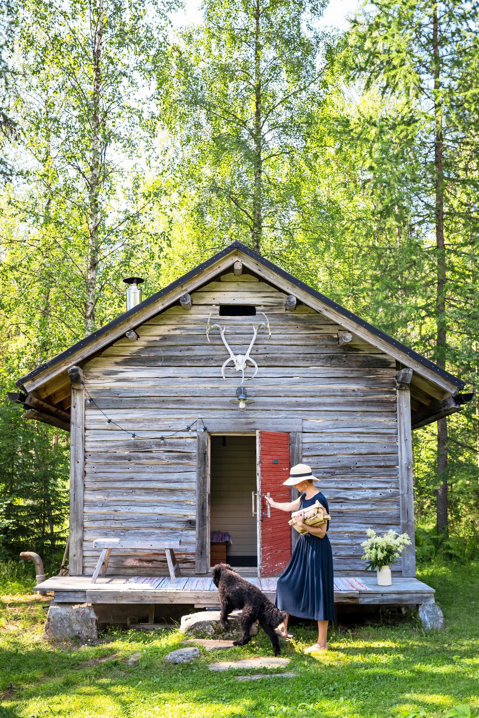 Nainen ja koira menevät harmaantuneeseen hirsirakennukseen.