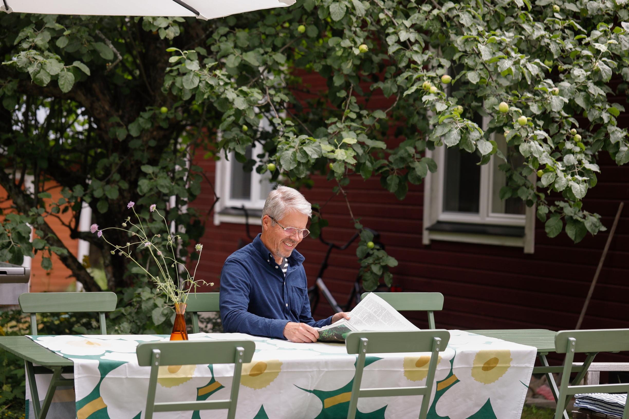 Hymyilevä mies lukee sanomalehteä kuuden hengen puutarharyhmässä, taustalla punainen talo, Unikko-pöytäliina.