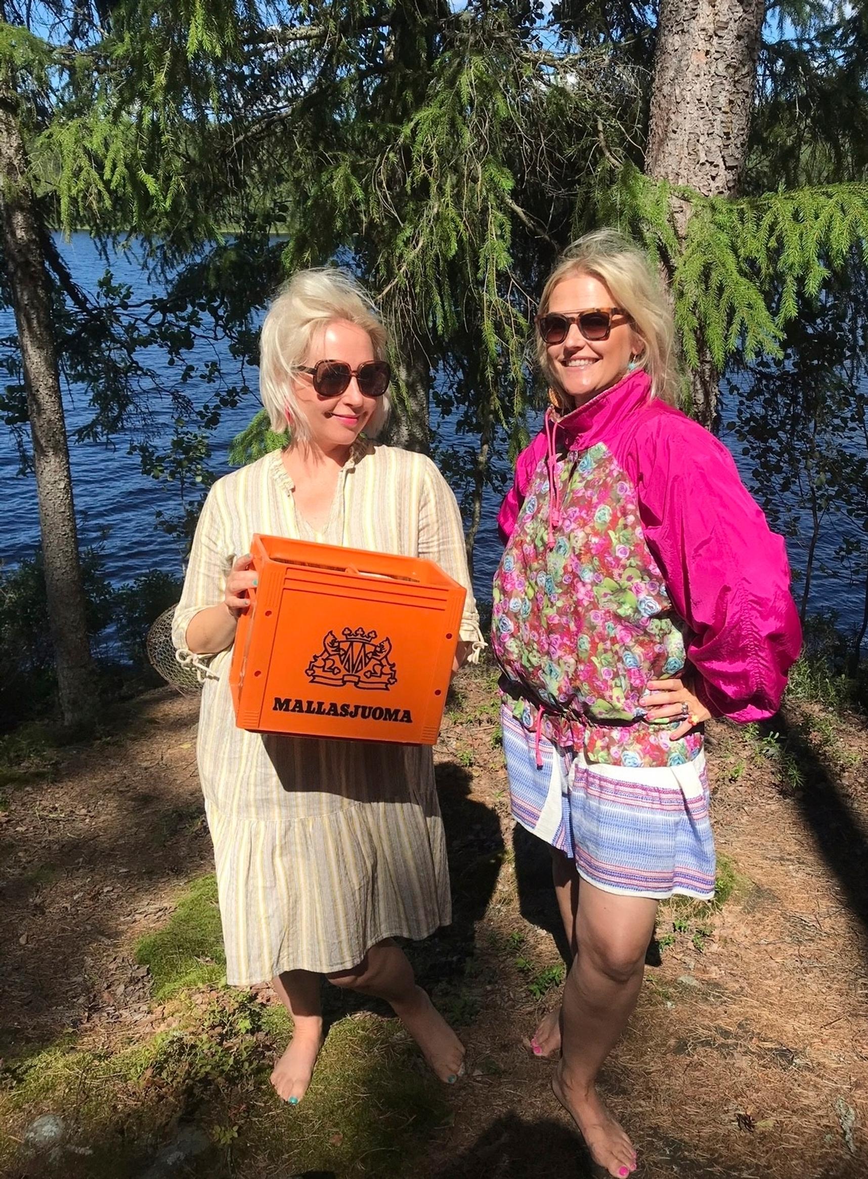 Näyttelijä Krisse Salminen toisen naisen kanssa näyttää vanhaa oranssia kaljakoria, molemmilla aurinkolasit, taustalla järvi