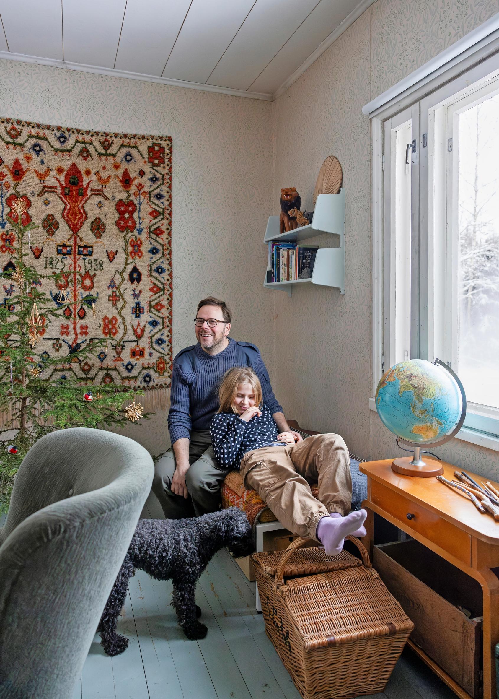Tytär Elsa ja isä Klaus Aalto mökillä Kiskossa sohvalla, taustalla ryijy, vieressä koira, ikkunalla karttapallo