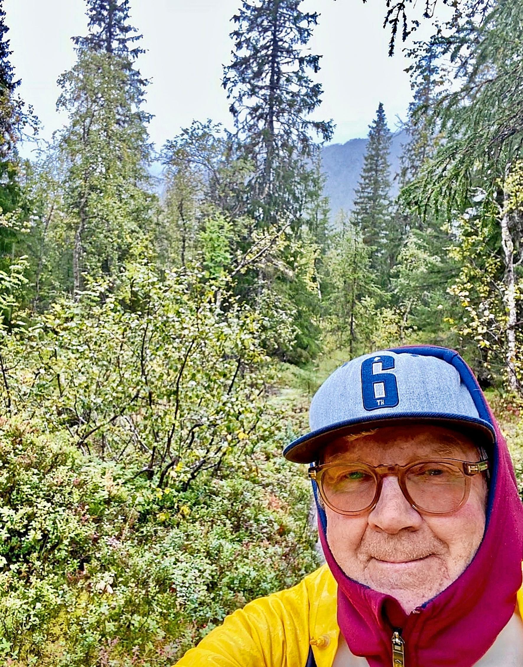 Pirkka-Pekka Petelius lippis ja huppari päässä mökkipuutarhassa, taustalla metsää