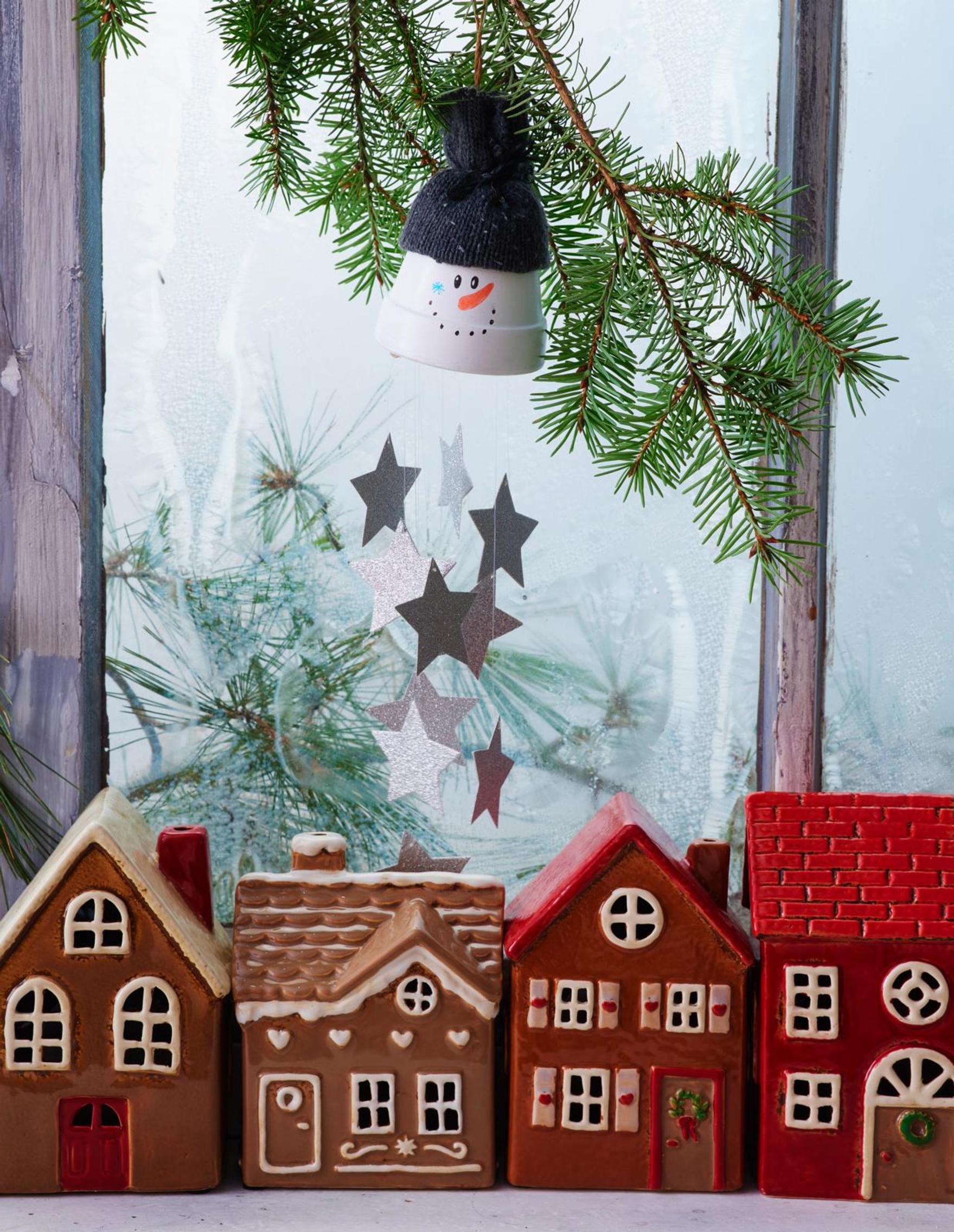 Valkoiseksi maalattu ruukku, jolle on piirretty silmät, suu ja oranssi nenä tussilla. Ruukun sisältä roikkuu hopeisia koristetähtiä siimassa. Lumiukolle on kääräisty kankaasta myssy ja se roikkuu havuoksasta. Alapuolella on talolyhtyjä.