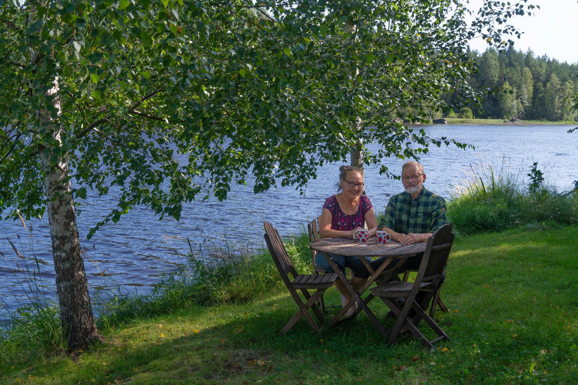 Riitta ja Pekka Vaaramo istuvat pyöreän puutarhapöydän ääressä rantakoivun vieressä nurmikolla, taustalla järveä ja vastaranta