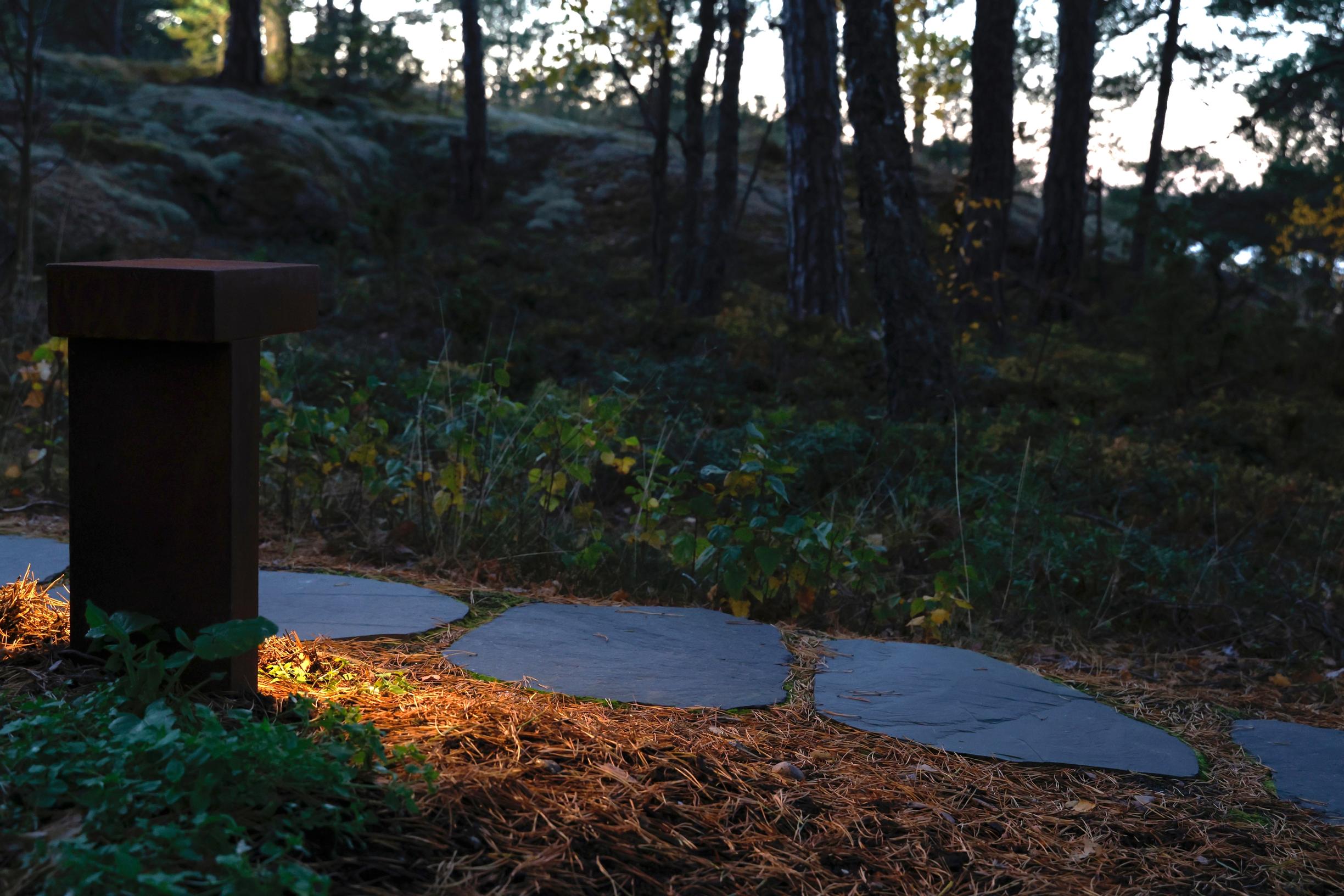 liuskekivillä peitetty polku ja tolppamainen ulkovalaisin, taustalla metsää ja meri