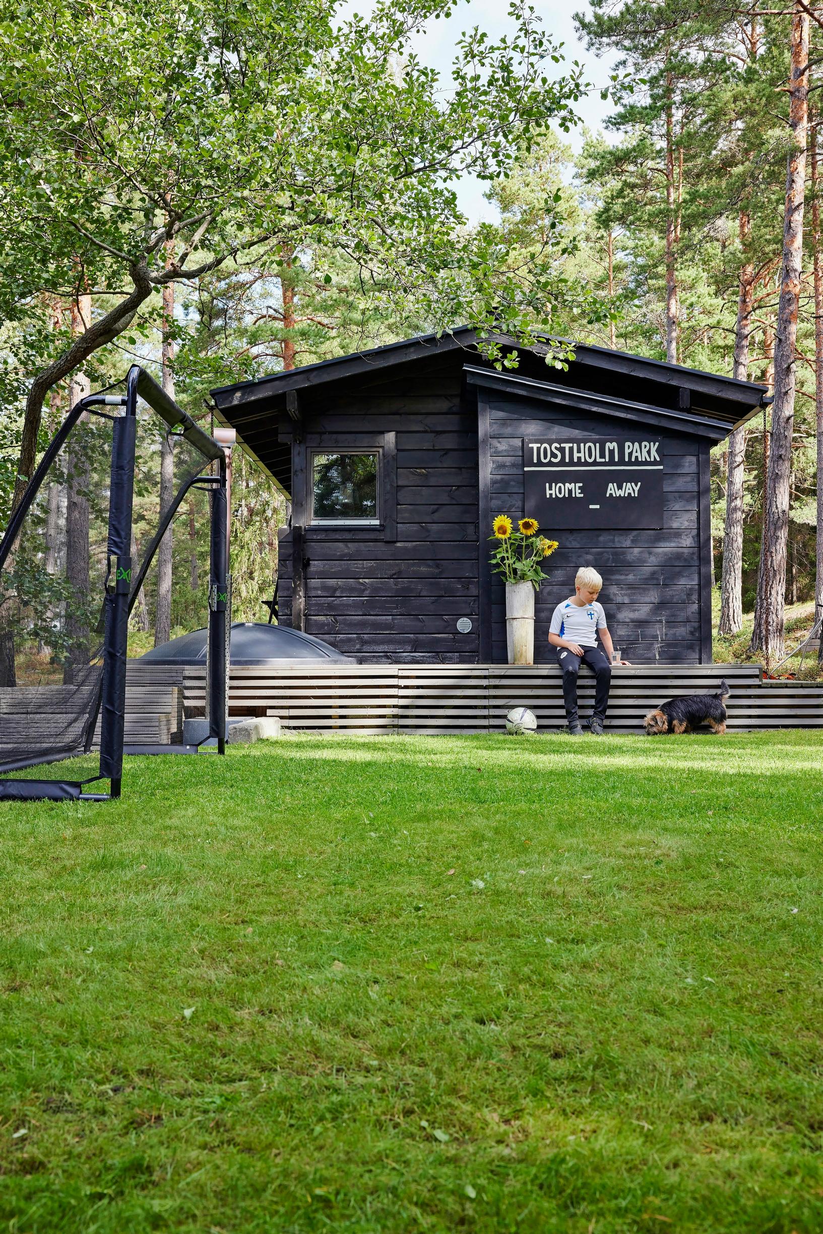 tumma sauna, jonka terassilla istuu poika, edessä koira vihreällä nurmikentällä ja jalkapallomaali